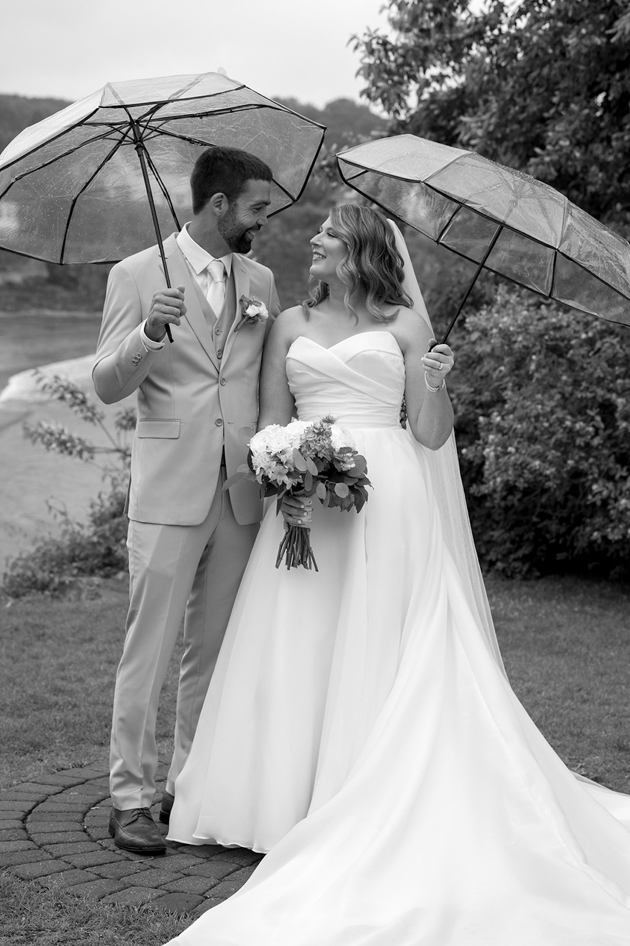 Newlyweds walking outdoors holding clear umbrellas during a rainy wedding portrait session.