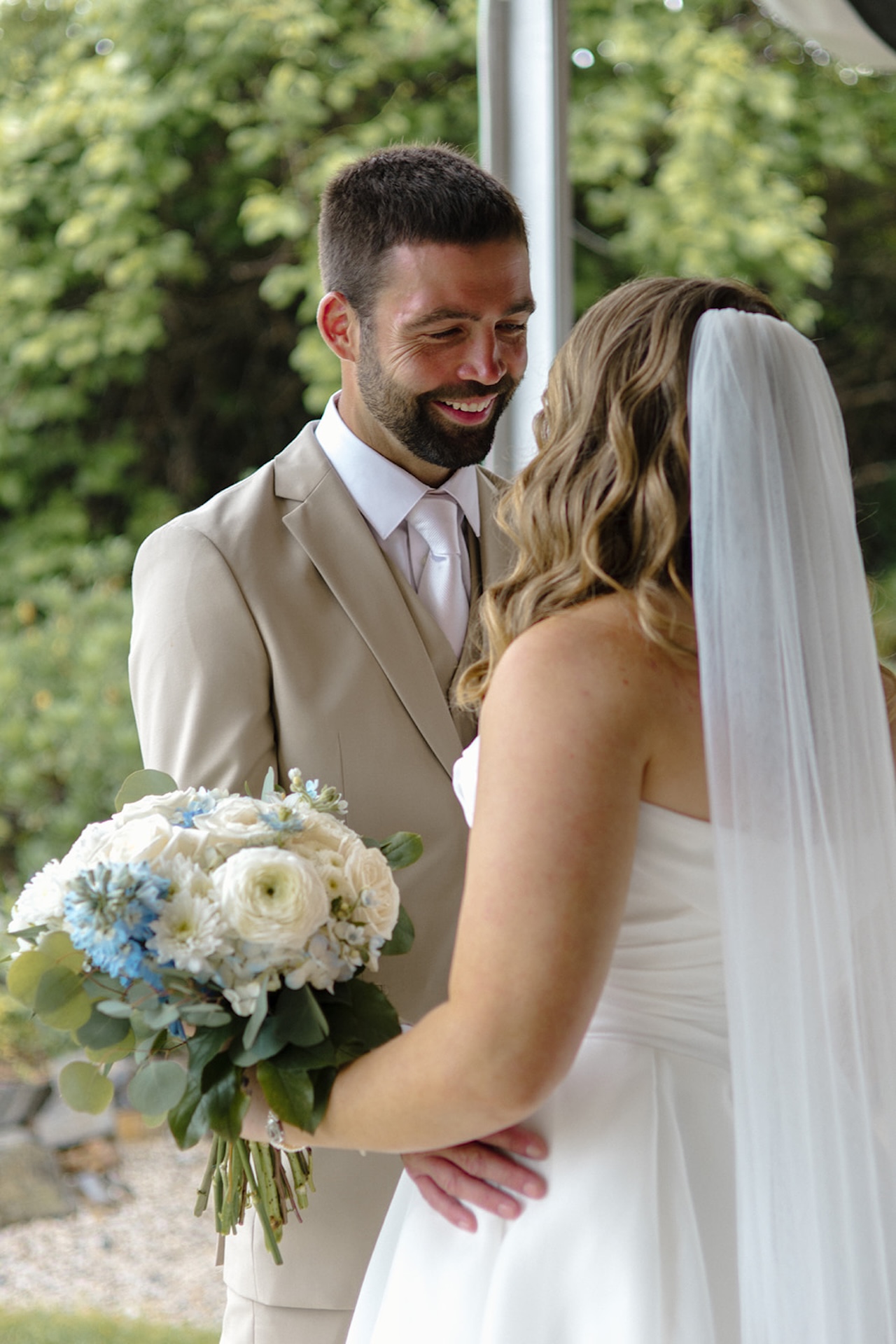 Bride and groom smiling during their first look while holding a blue and white floral bouquet outdoors.