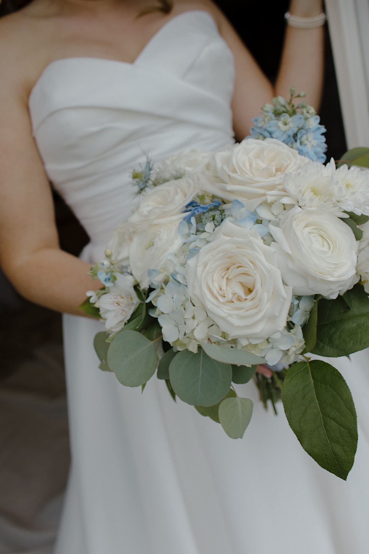 Bride standing in a doorway holding a white and blue coastal wedding bouquet, looking out with soft natural light.