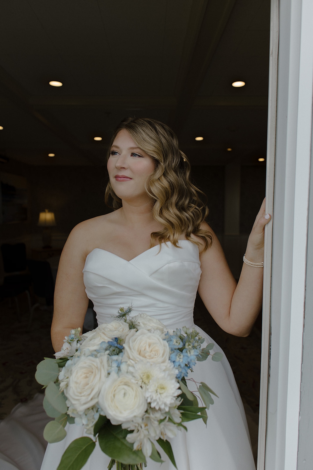 Bride standing in a doorway holding a white and blue coastal wedding bouquet, looking out with soft natural light.