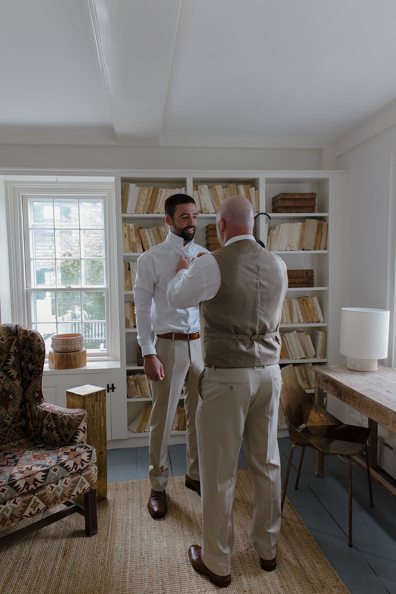 Groom getting ready while a groomsman adjusts his tie in a bright room with neutral coastal decor.
