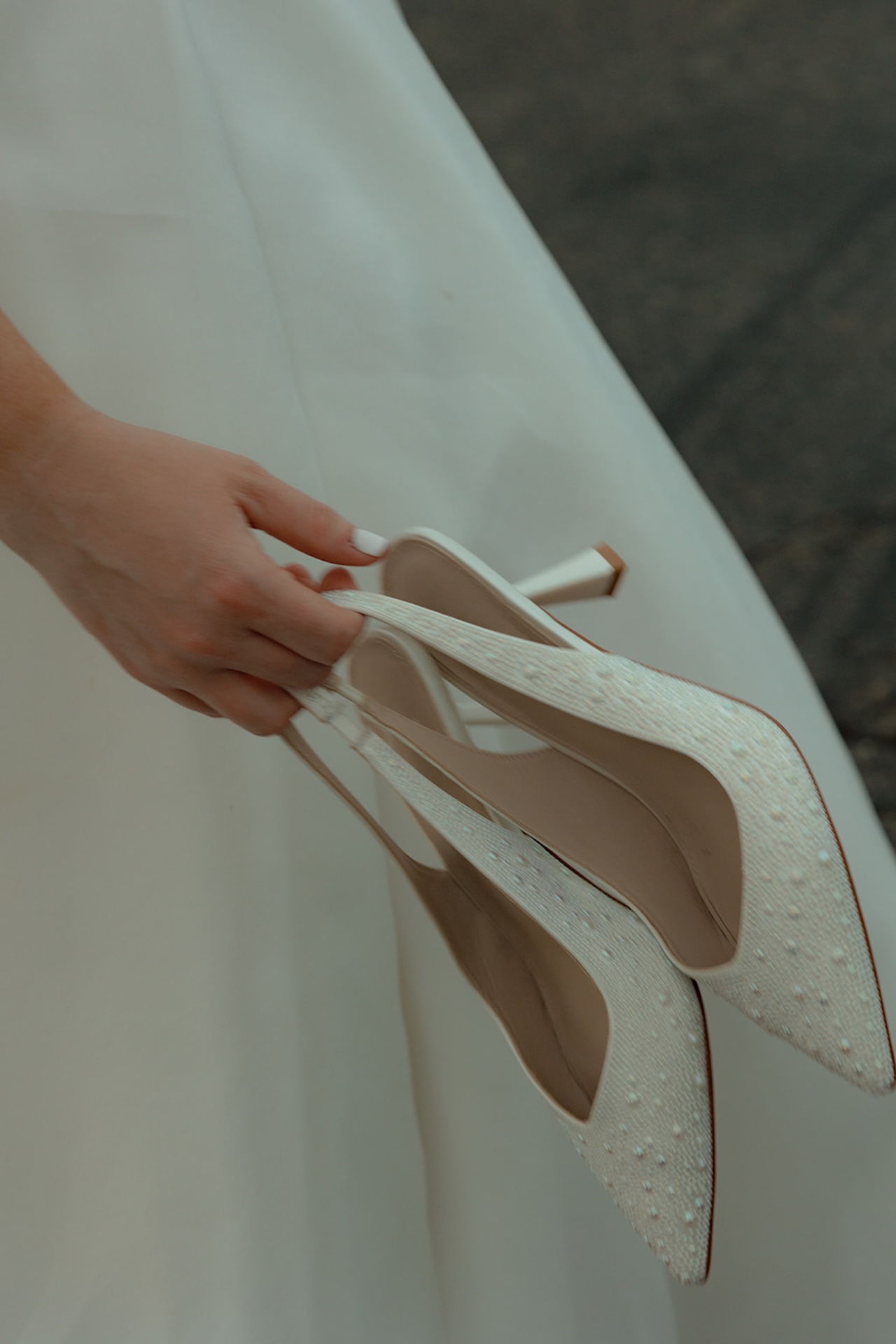 Bride holding pearl embellished wedding heels against the backdrop of her wedding dress.