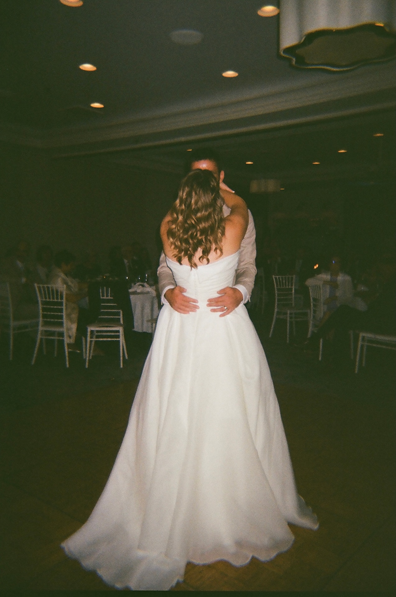 Newlyweds sharing their first dance in a dimly lit reception space surrounded by guests.