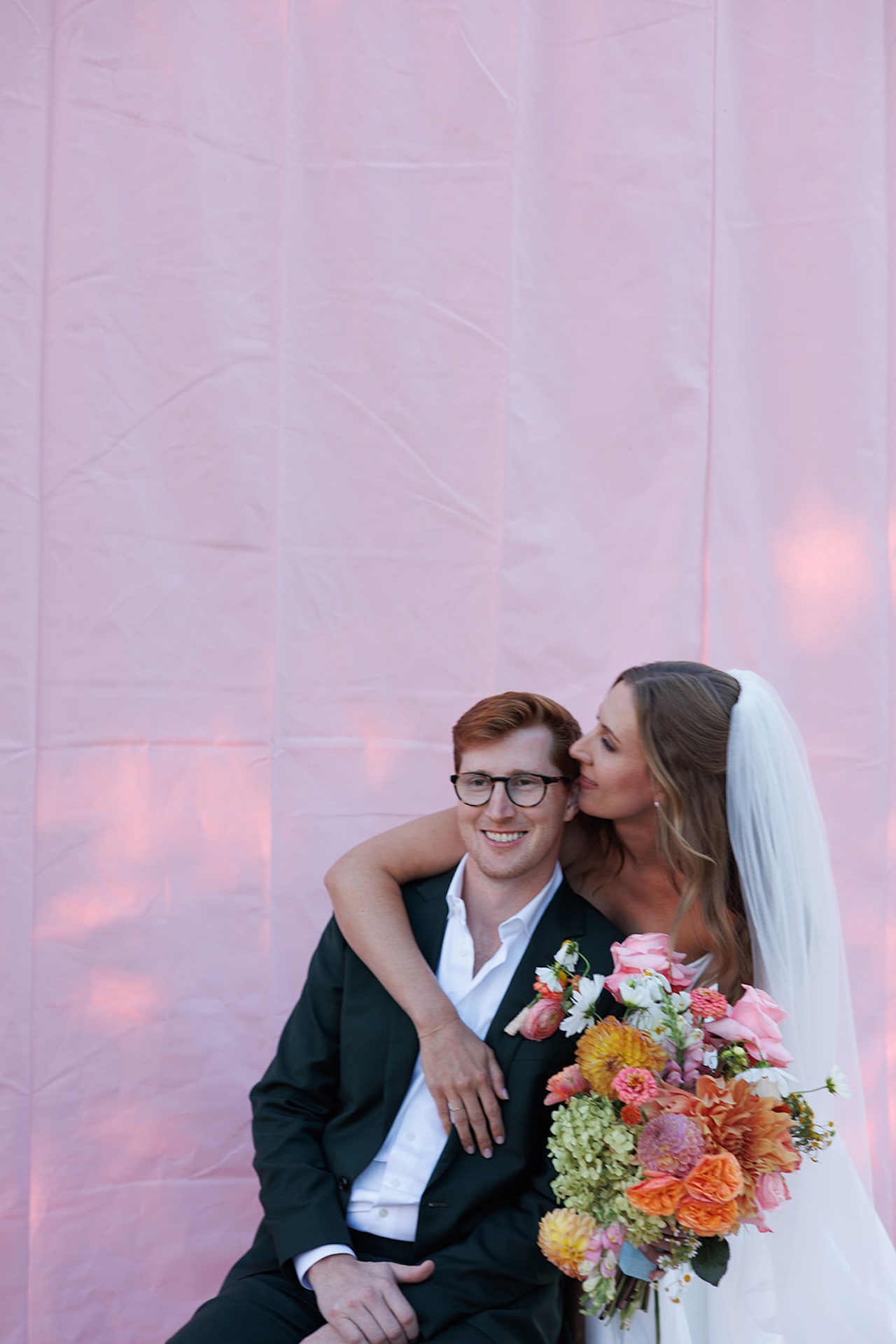Bride and groom portraits with the bride holding a colorful bouquet with bright pinks, oranges, yellows, and purples with a bright pink backdrop behind them. Perfect inspiration for colorful weddings.