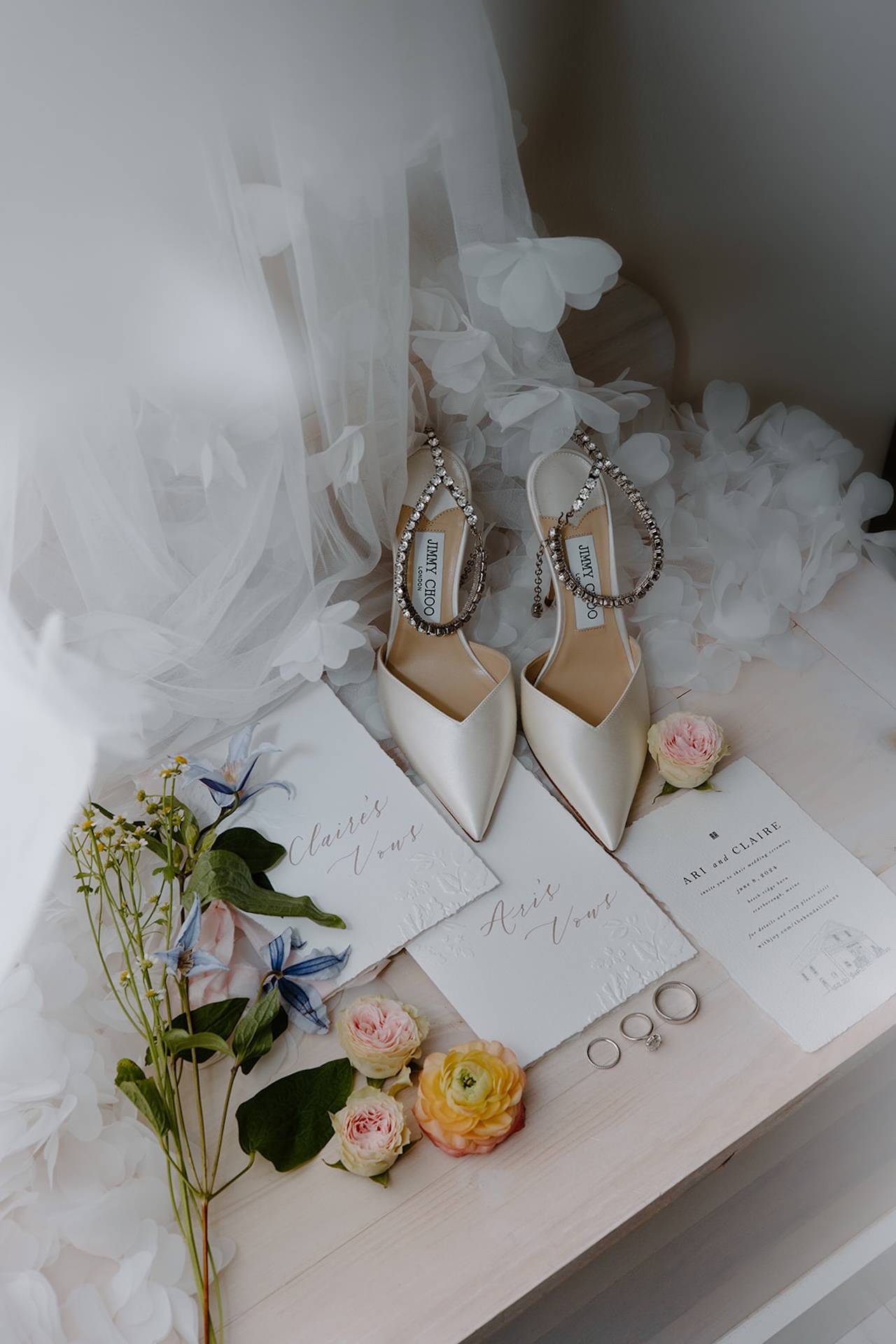 A wedding flat lay with white heels, a wedding invite, bright florals and the rings.