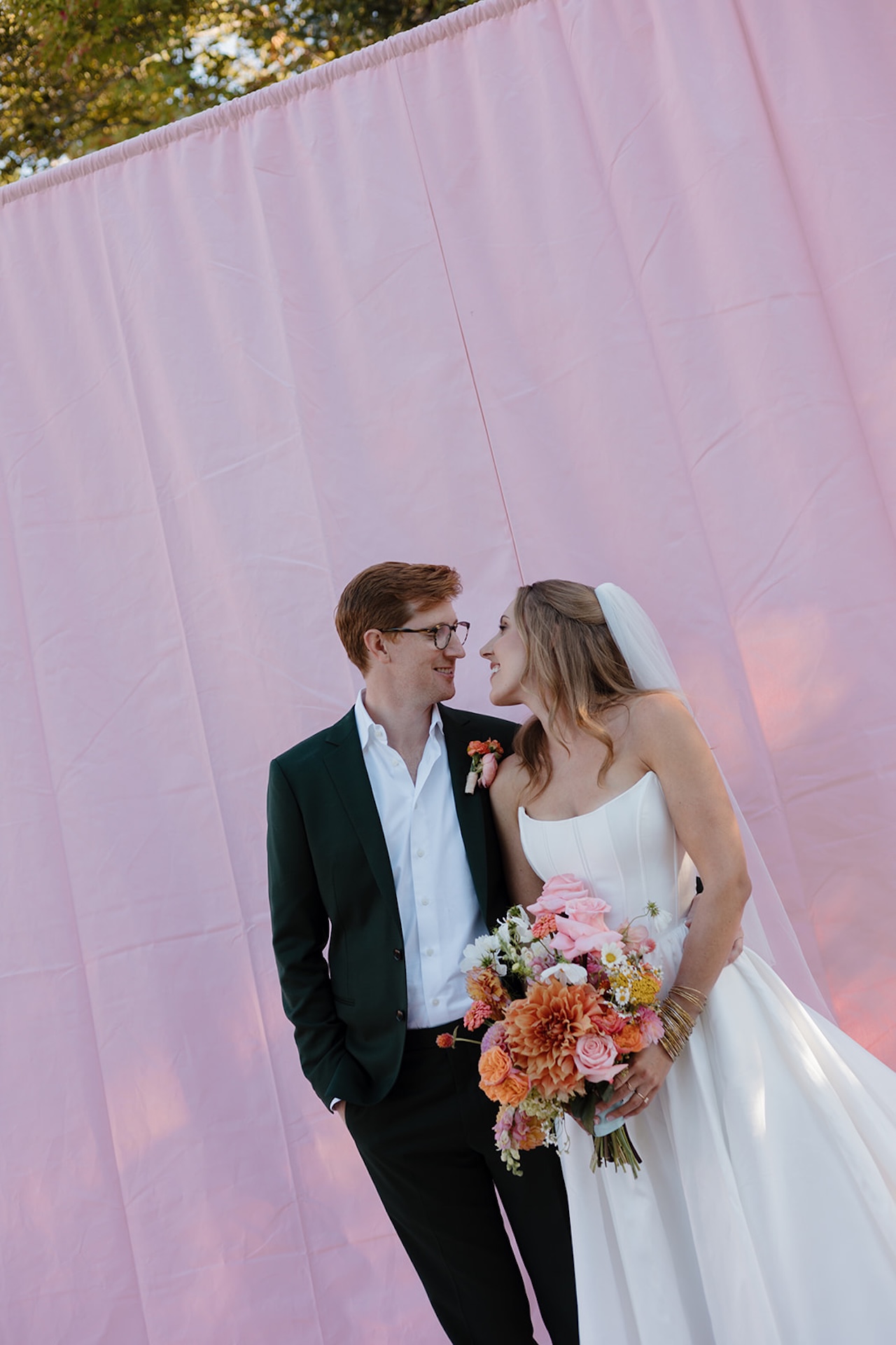 Bride and groom portraits with the bride holding a colorful bouquet with bright pinks, oranges, yellows, and purples with a bright pink backdrop behind them. Perfect inspiration for colorful weddings.