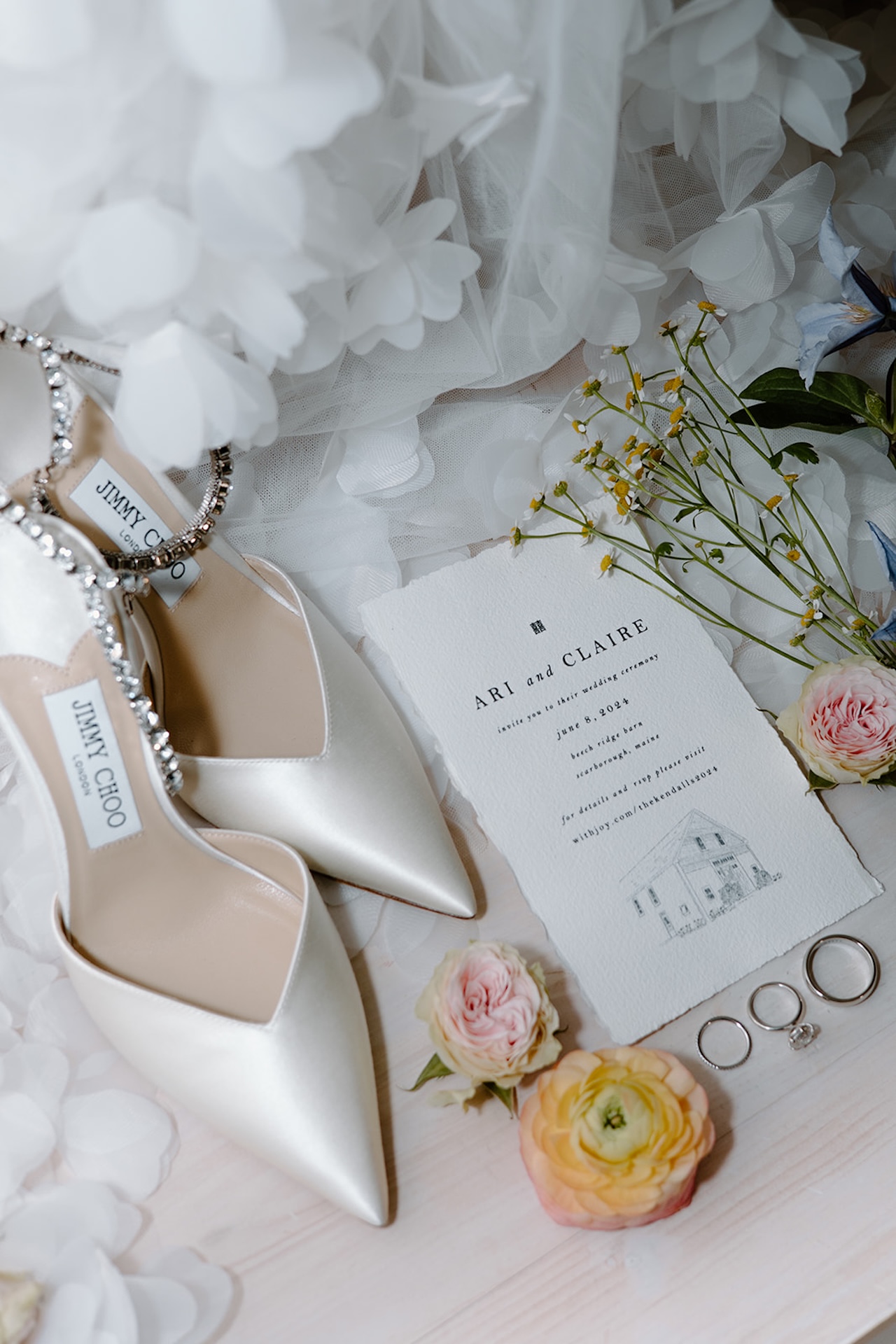 A wedding flat lay with white heels, a wedding invite, bright florals and the rings.
