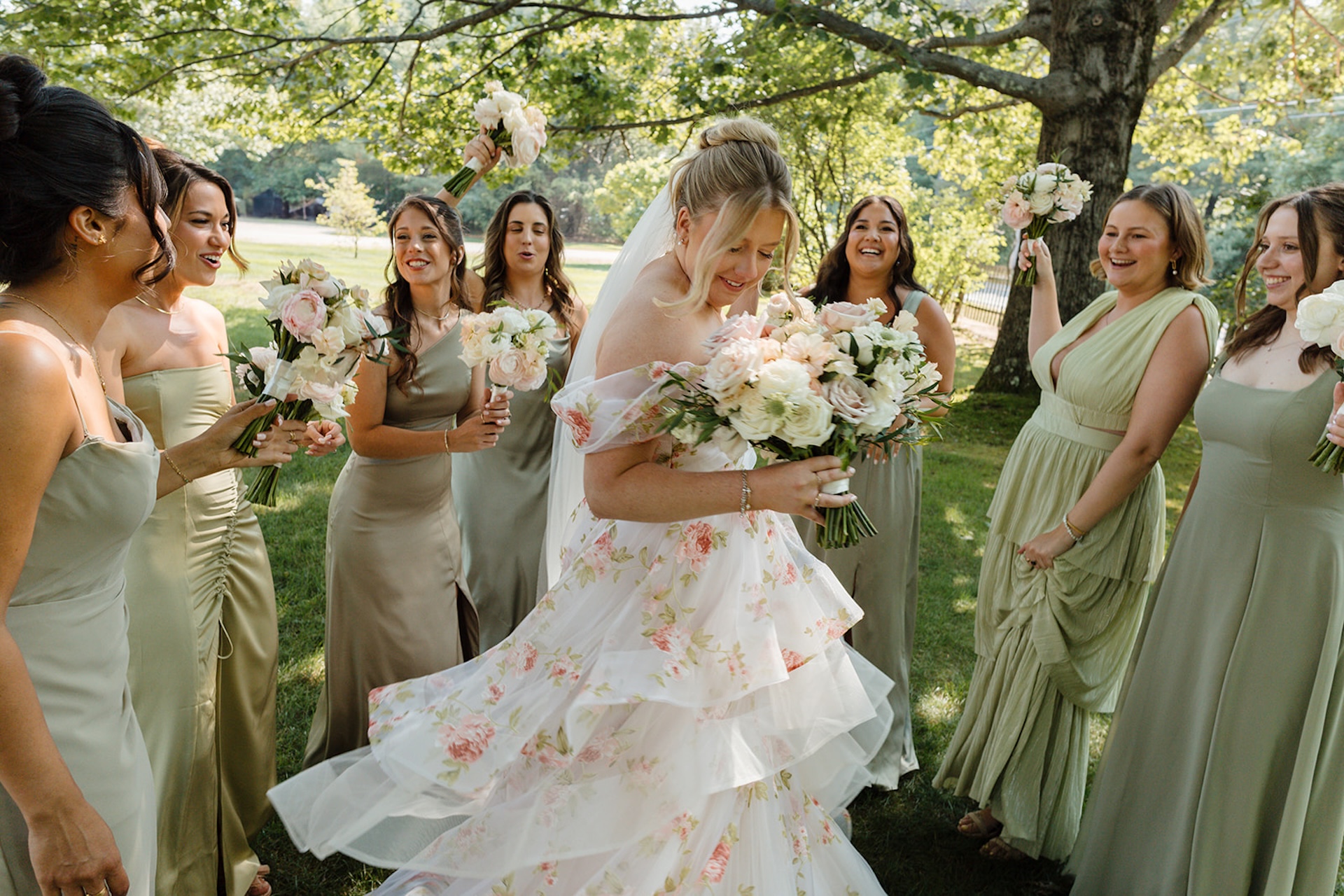 Bride in a floral white wedding dress spinning with a circle of her bridesmaids around her in green dresses and holding white and light pink florals.