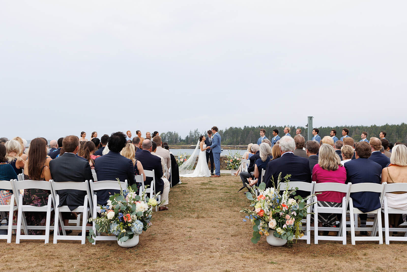 Wide view of a waterfront micro wedding ceremony in coastal Maine with floral arrangements lining the aisle.