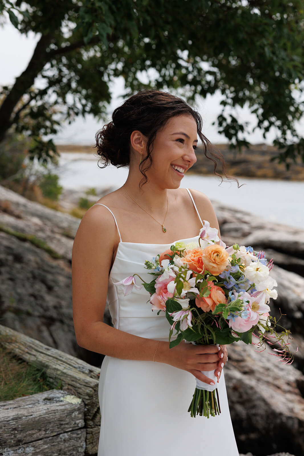 Bride smiling with colorful bouquet during a coastal Maine micro wedding beside rocky waterfront scenery.
