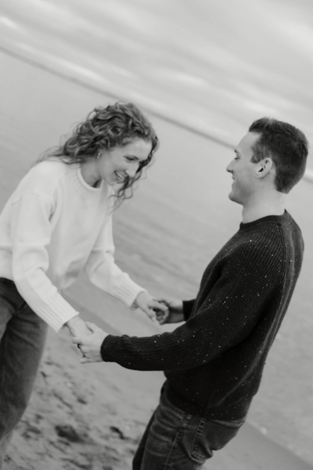 Black and white engagement photoshoot image of a couple laughing together on the beach