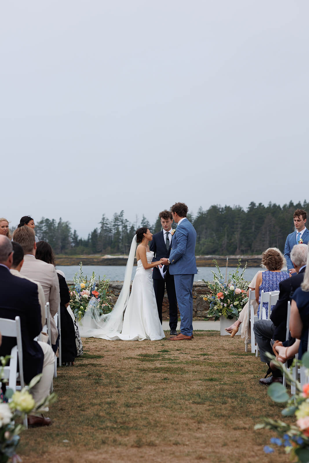 Intimate coastal Maine micro wedding ceremony overlooking the water with guests seated beside the bride and groom during their vows.