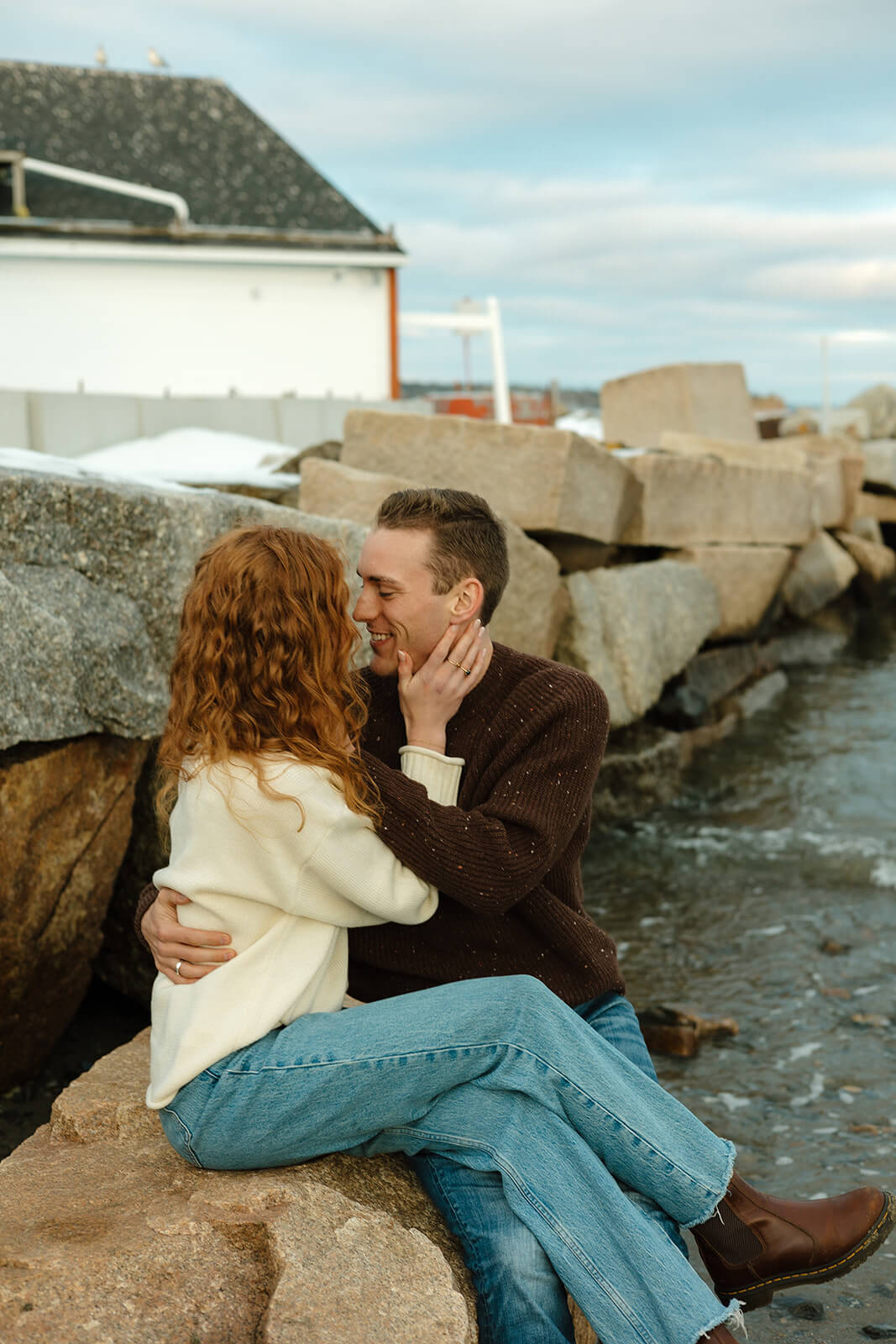 Couple cuddling on seaside rocks during a cozy engagement photoshoot in Maine