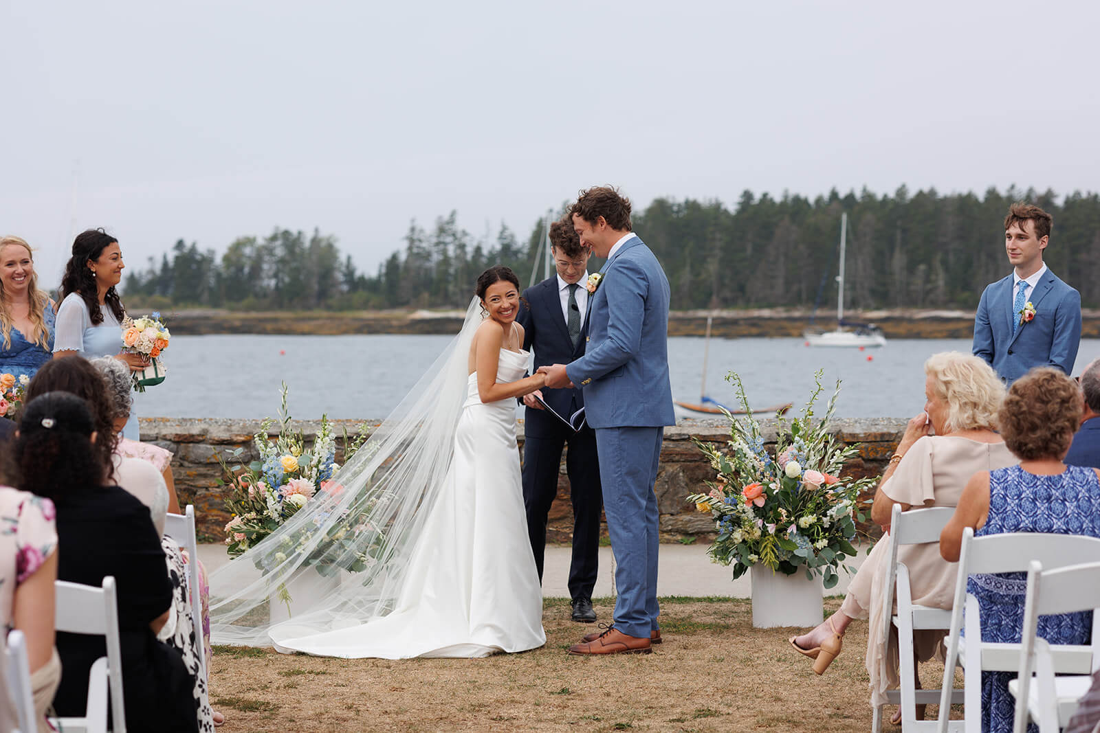 Bride smiling during a waterfront micro wedding ceremony in coastal Maine surrounded by family and pastel floral arrangements.
