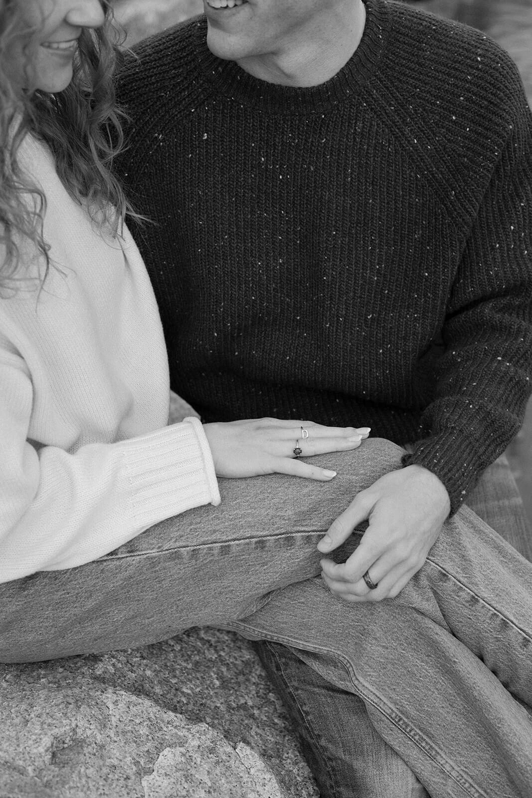 Black and white candid engagement photoshoot image of a couple sitting together on seaside rocks