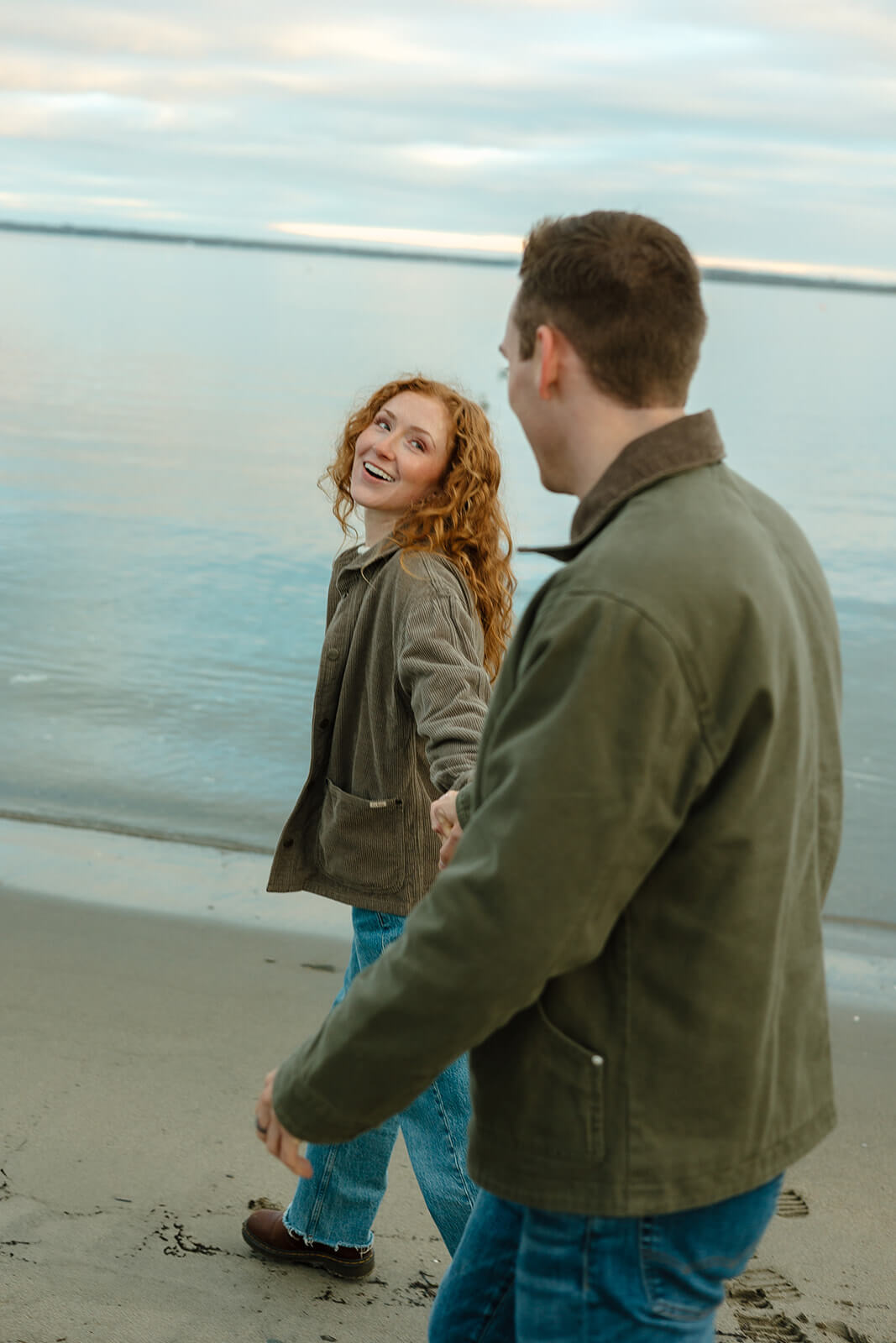 Couple walking hand-in-hand along the shoreline during a candid coastal engagement photoshoot