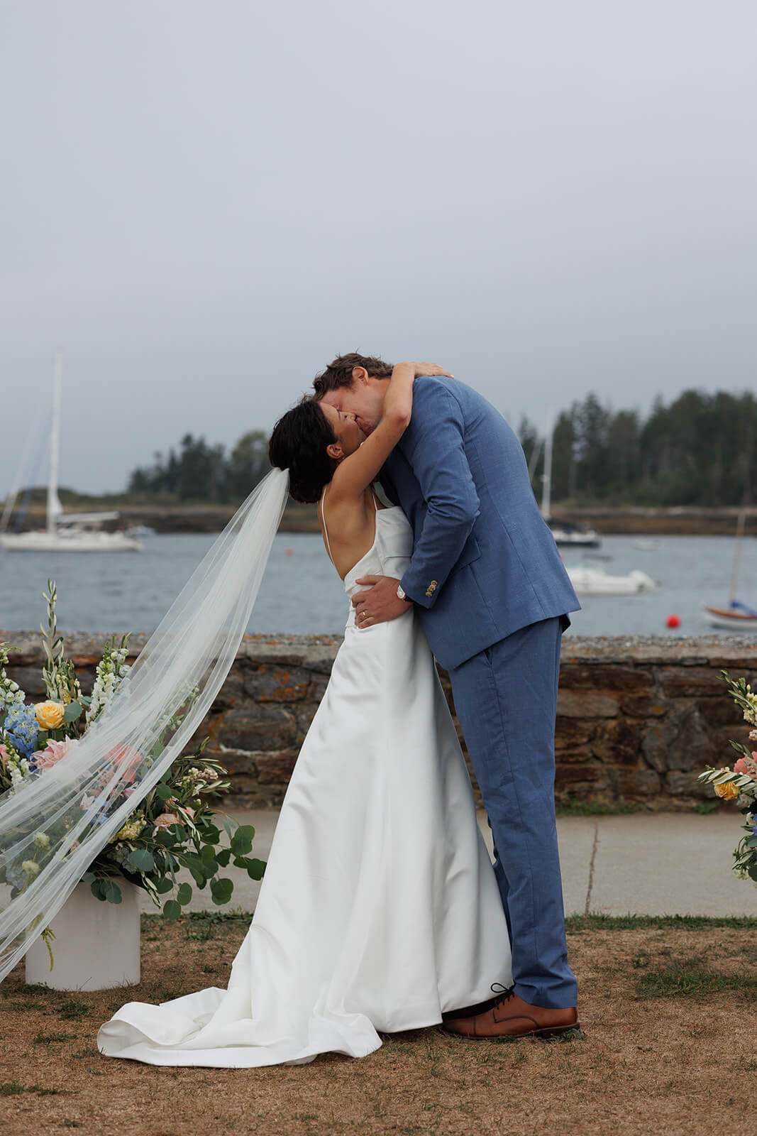 Newly married couple kissing during their coastal Maine micro wedding ceremony with sailboats and ocean views behind them.