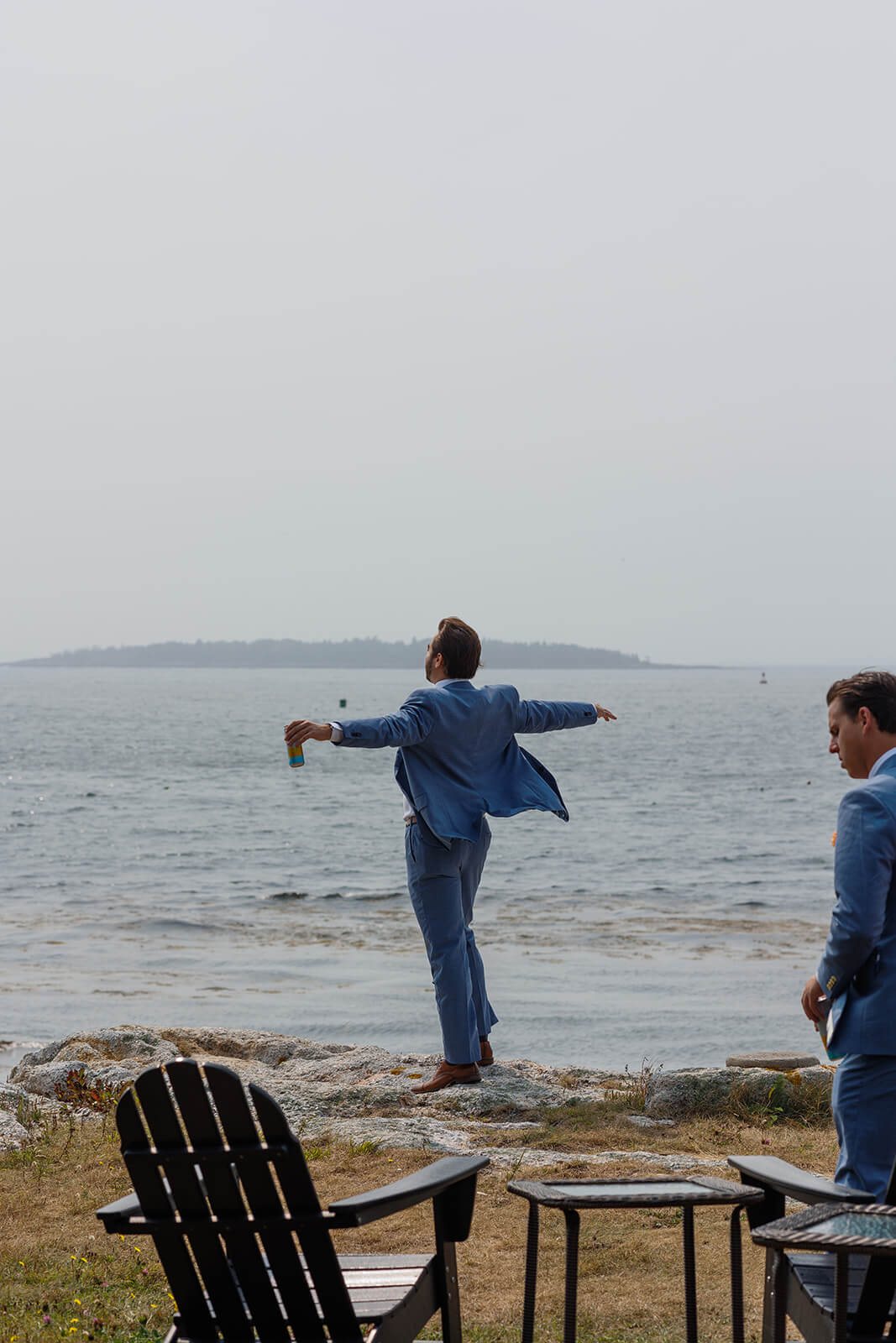 Groomsman in blue suit spinning along the rocky waterfront before a coastal Maine micro wedding.