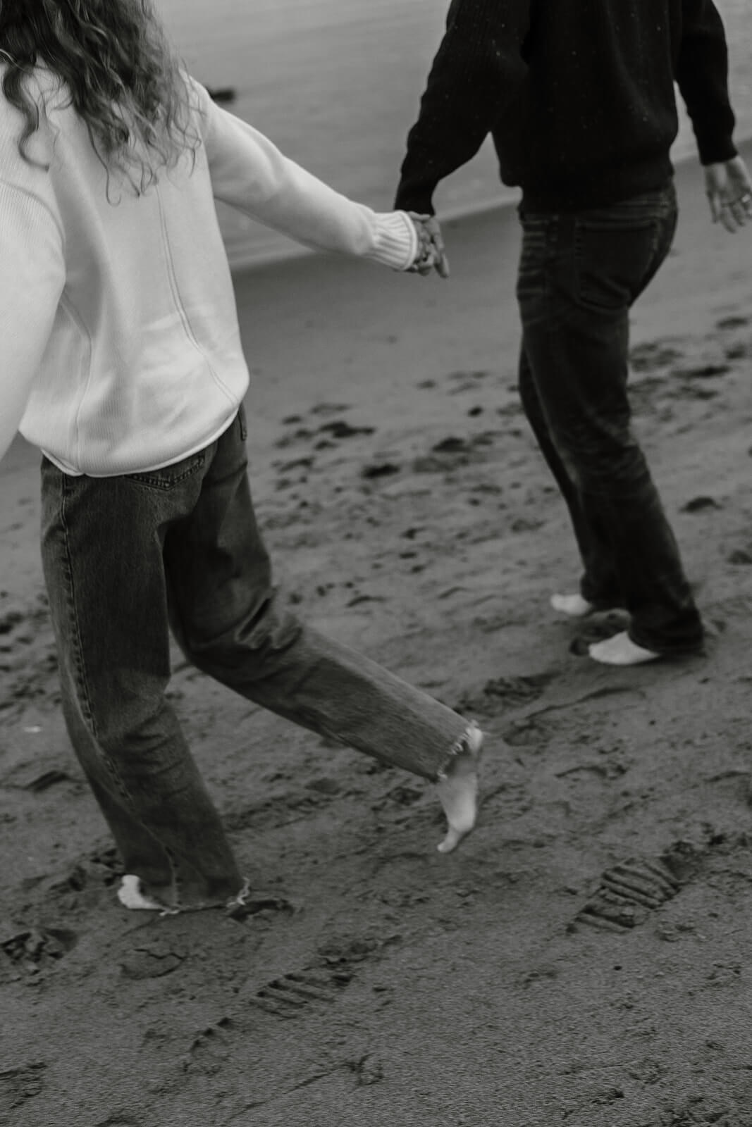 Black and white candid engagement photoshoot image of a couple walking barefoot along the shoreline