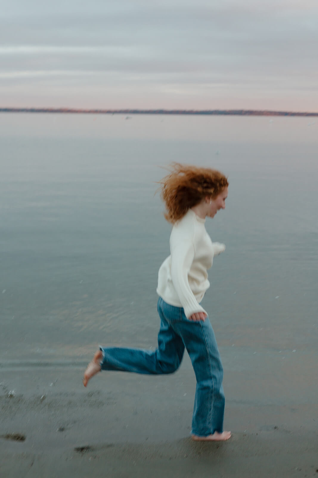 Playful engagement photoshoot image of a woman running barefoot along the shoreline during a coastal Maine beach session