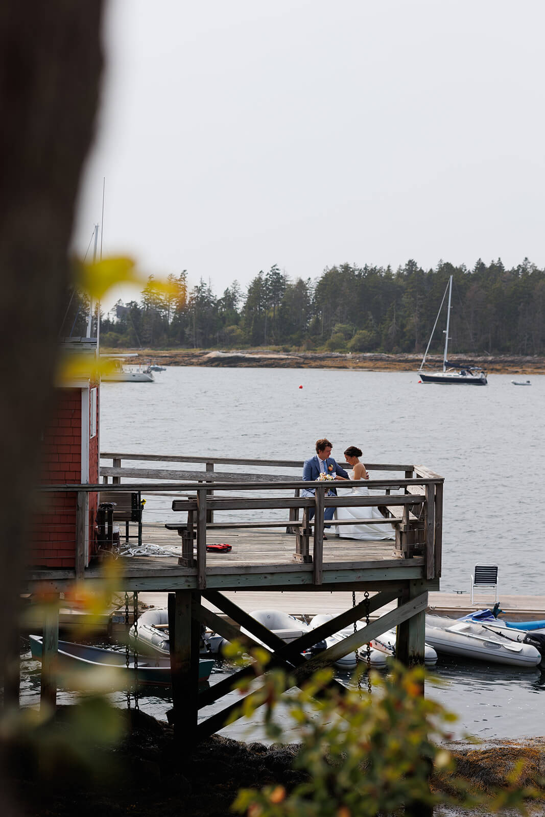 Bride and groom standing on a wooden dock surrounded by sailboats during a coastal Maine wedding portrait session.