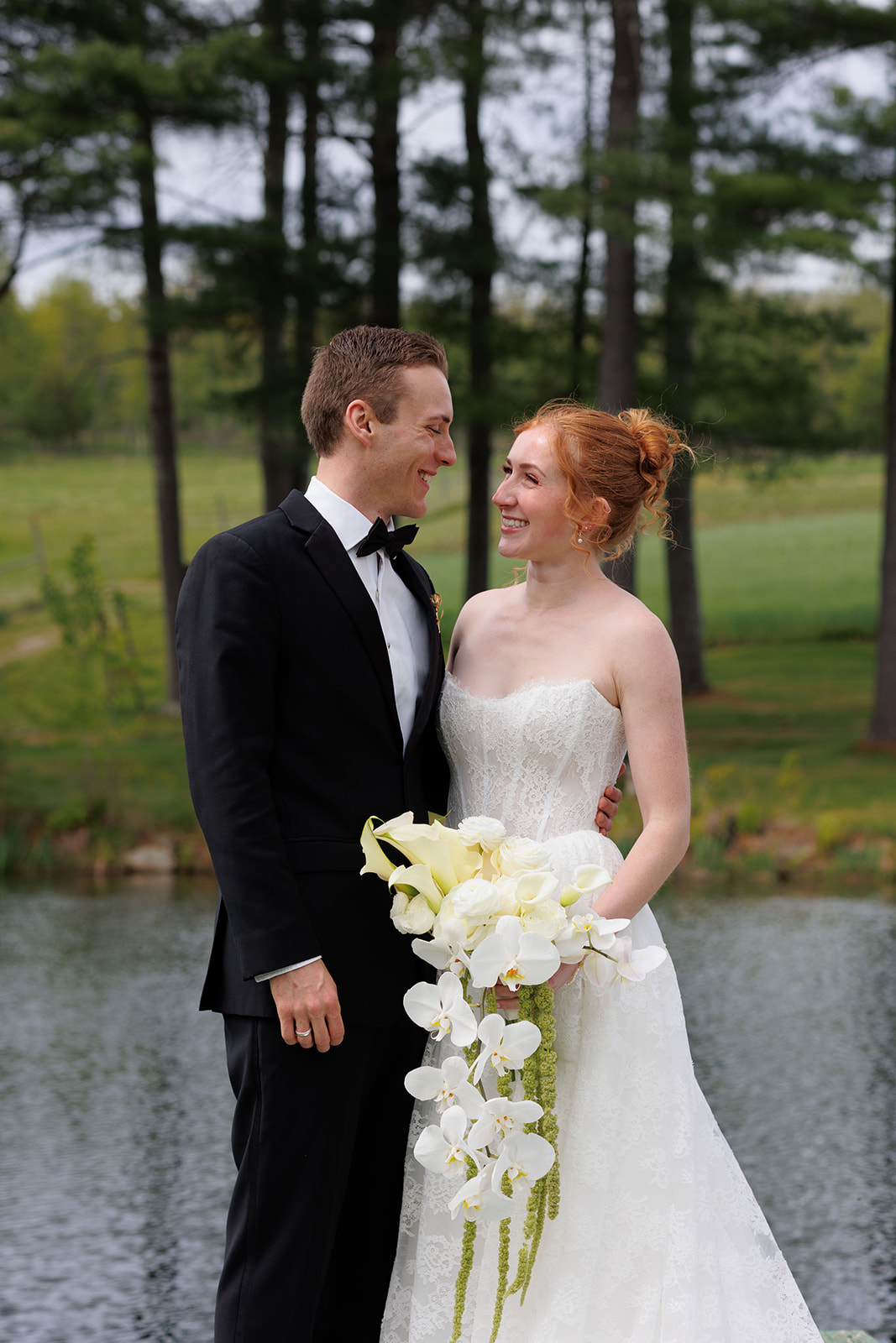 Bride and groom smiling at each other beside a pond, with the bride holding a cascading white orchid bouquet.