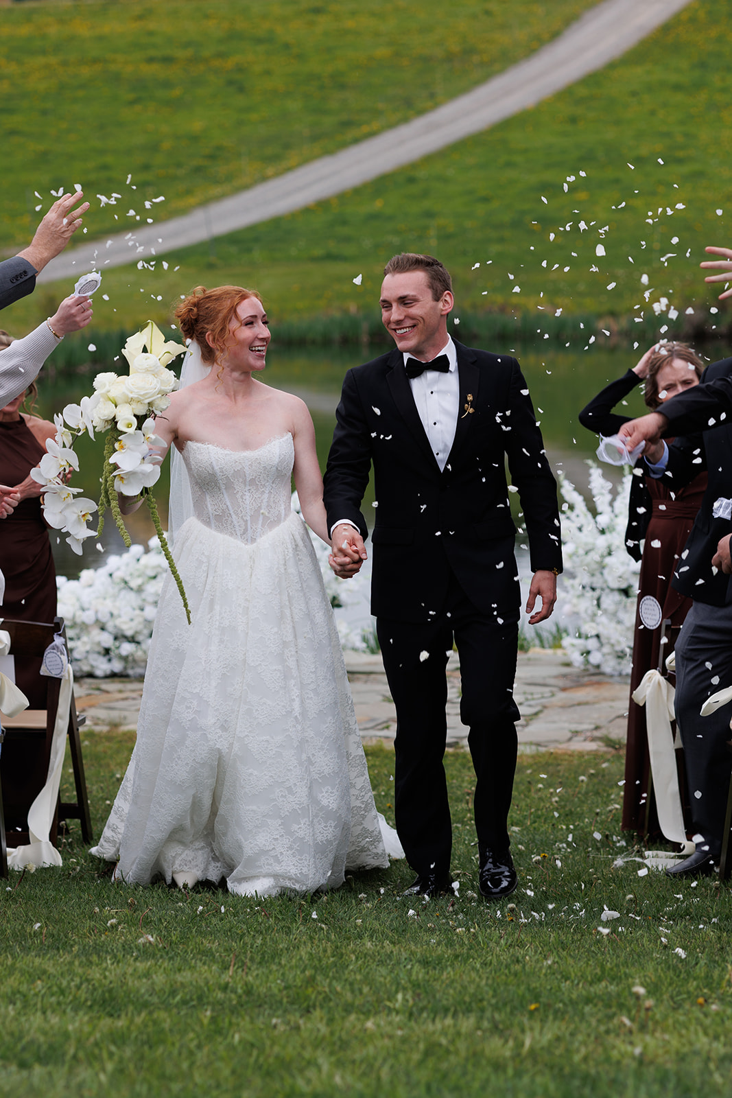 Newlyweds walking up the aisle hand in hand as guests toss white flower petals around them.