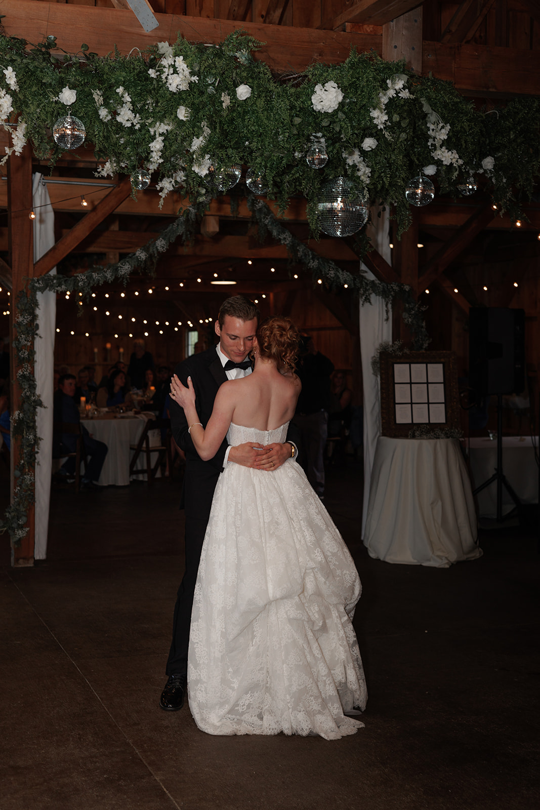 Newlyweds sharing their first dance inside a rustic barn decorated with greenery and string lights.