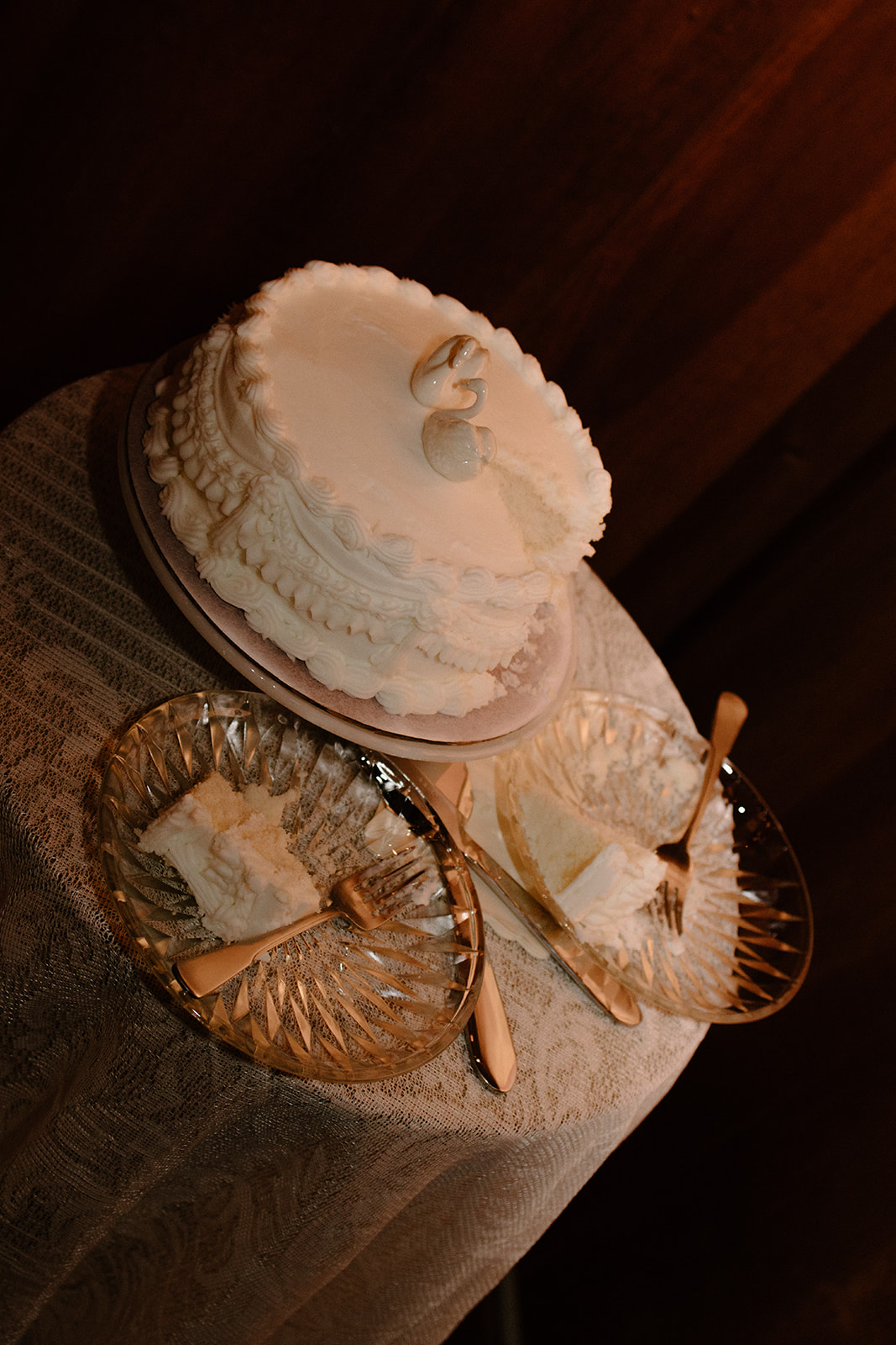 Small white wedding cake with decorative icing and intertwined rings displayed on a lace-covered table.