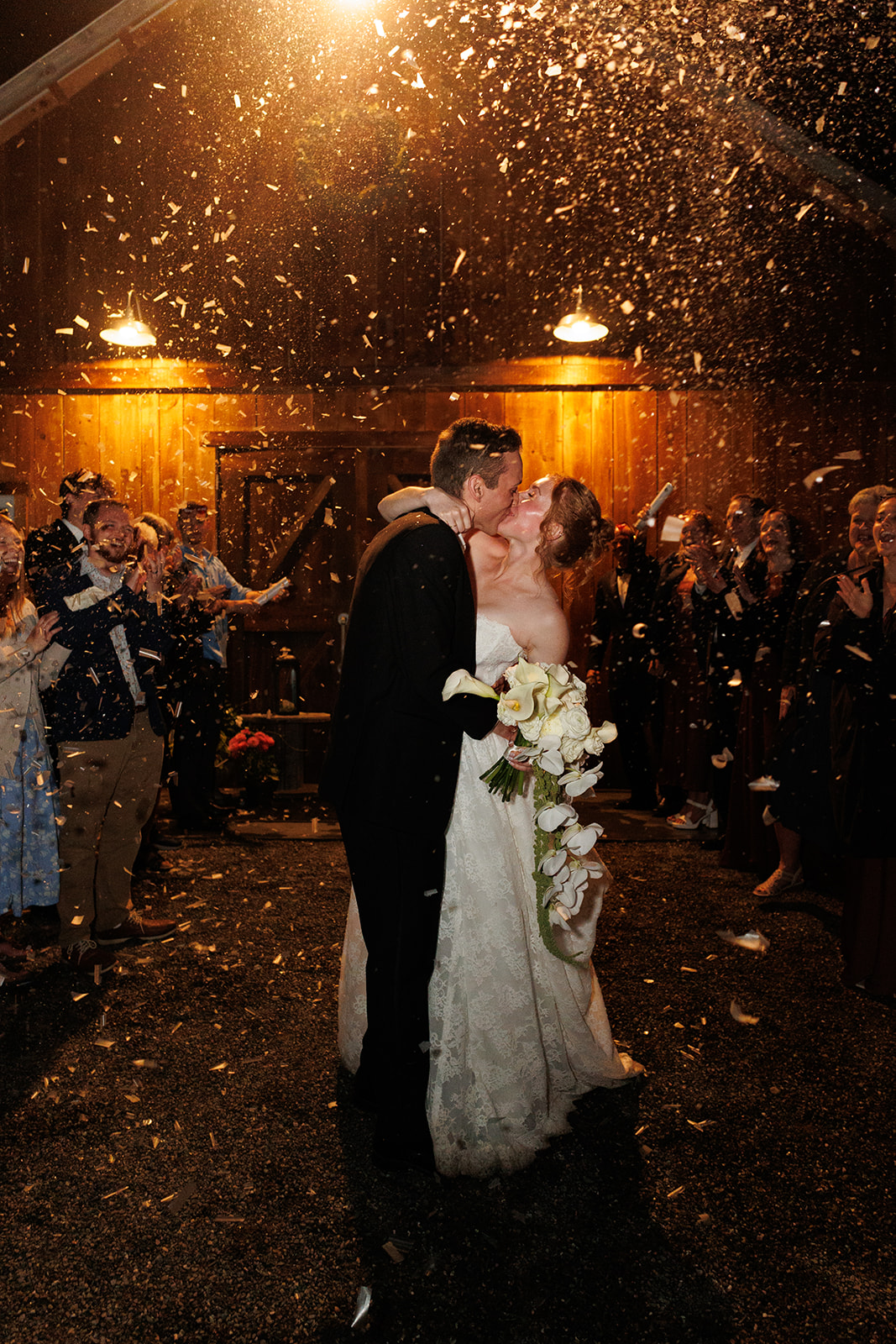Newlyweds kissing outside the barn at night while guests celebrate around them with sparkling confetti.