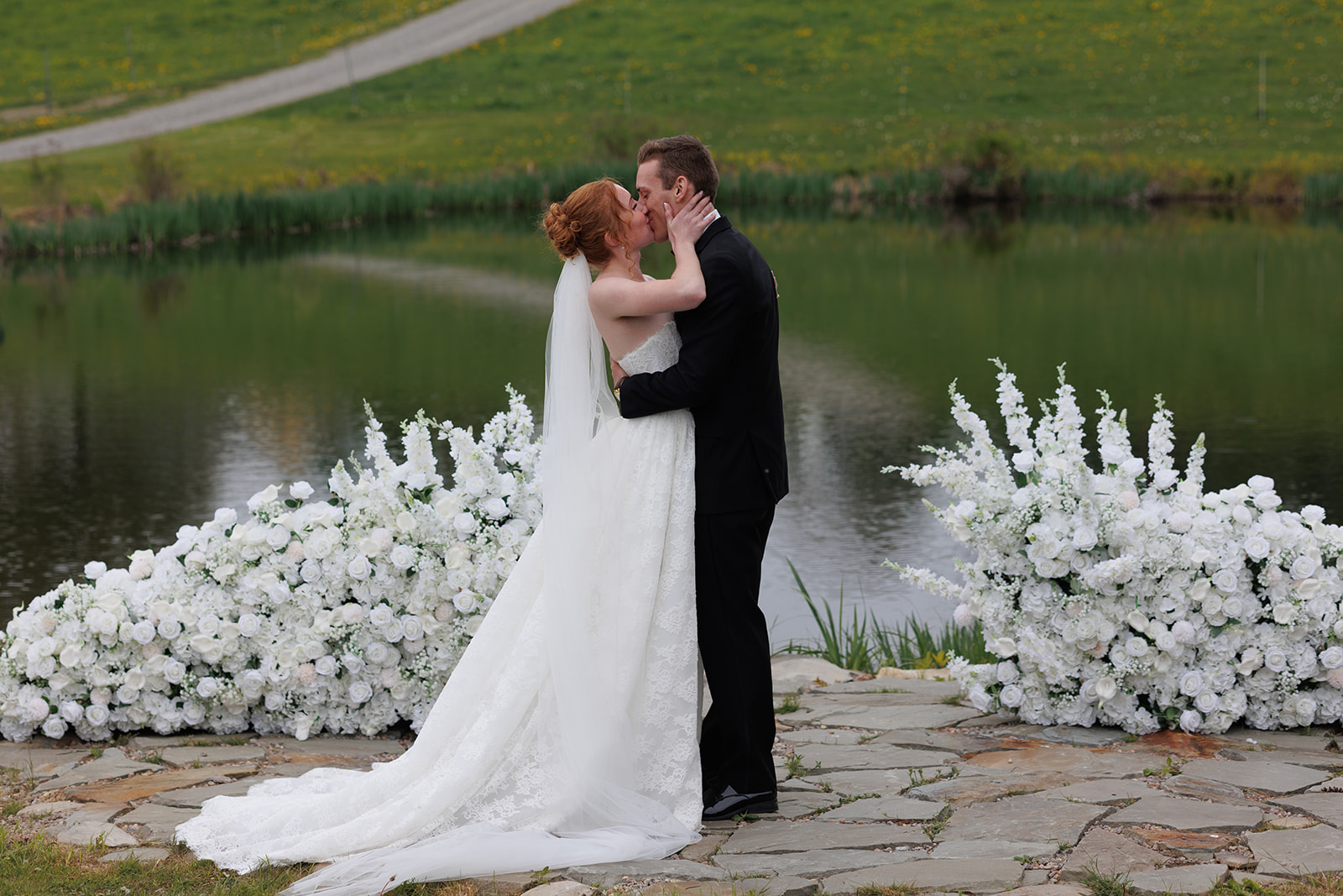 Newlyweds sharing their first kiss beside a pond, framed by overflowing arrangements of white flowers.