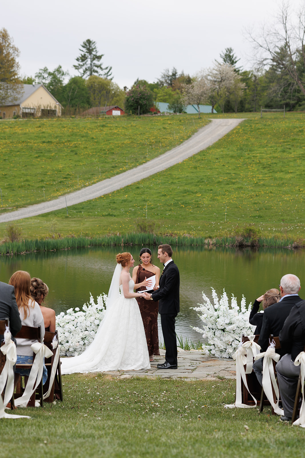 Outdoor wedding ceremony beside a pond with guests seated on the lawn and rolling hills in the distance.