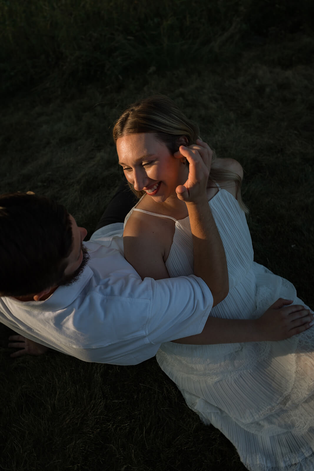 Overhead portrait of a couple embracing on the grass as the woman smiles and lifts a hand toward her hair.