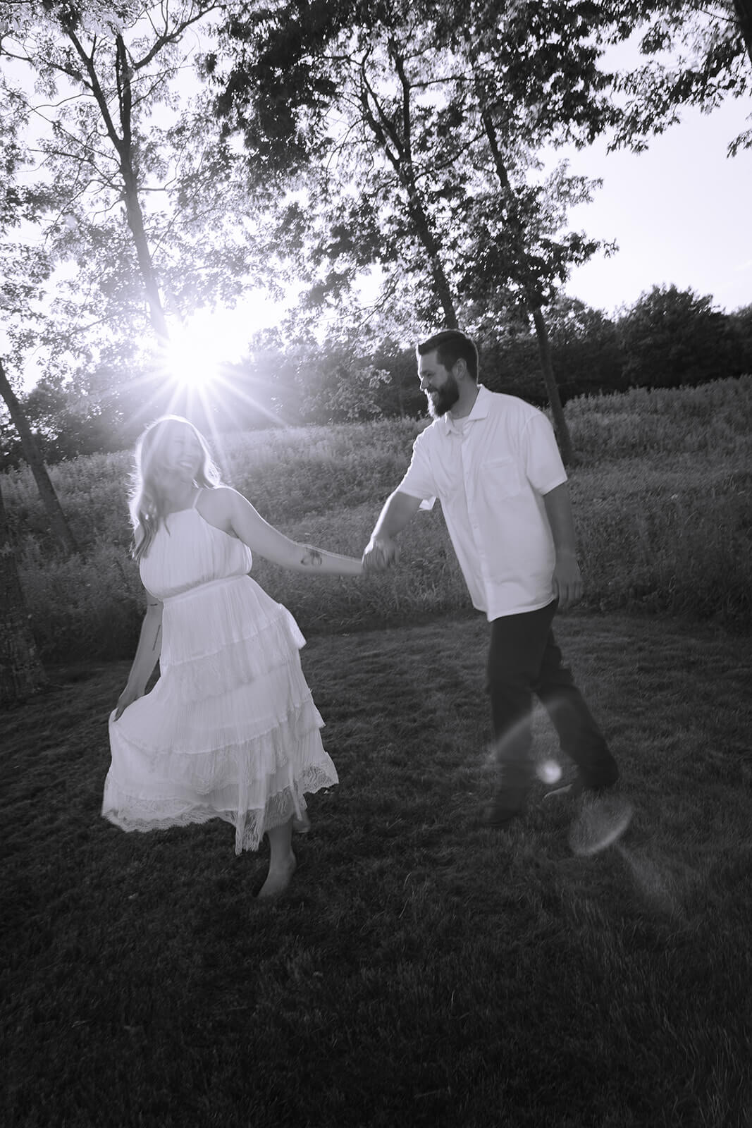 Black-and-white photograph of a couple walking and playfully moving hand in hand across the lawn with sunlight streaming through the trees at Beech Hill Barn.