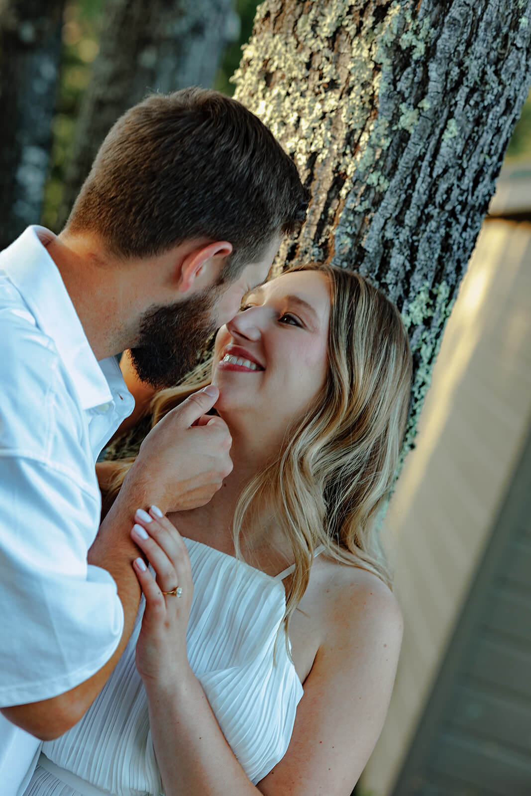 Man gently touching his partner’s chin as she smiles up at him beside a tree.