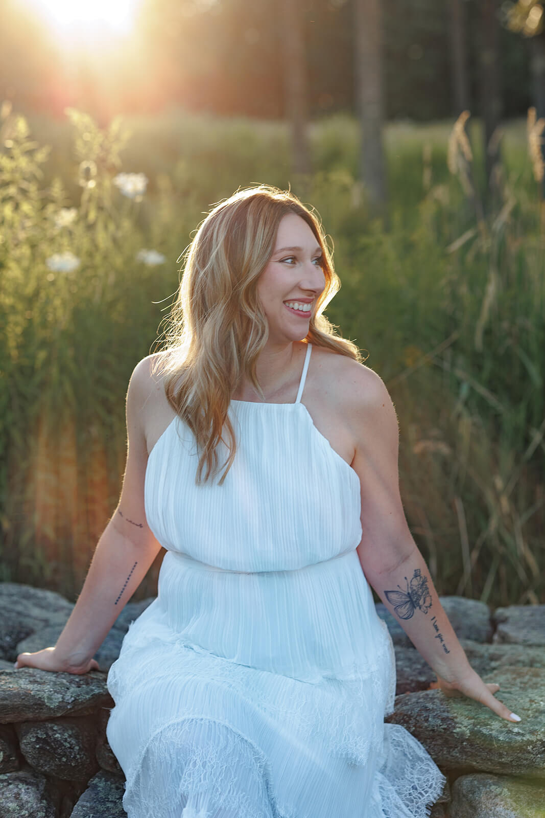 Woman in a white dress sitting on a stone wall and smiling to the side with warm sunlight behind her and tattoos visible on her arms.