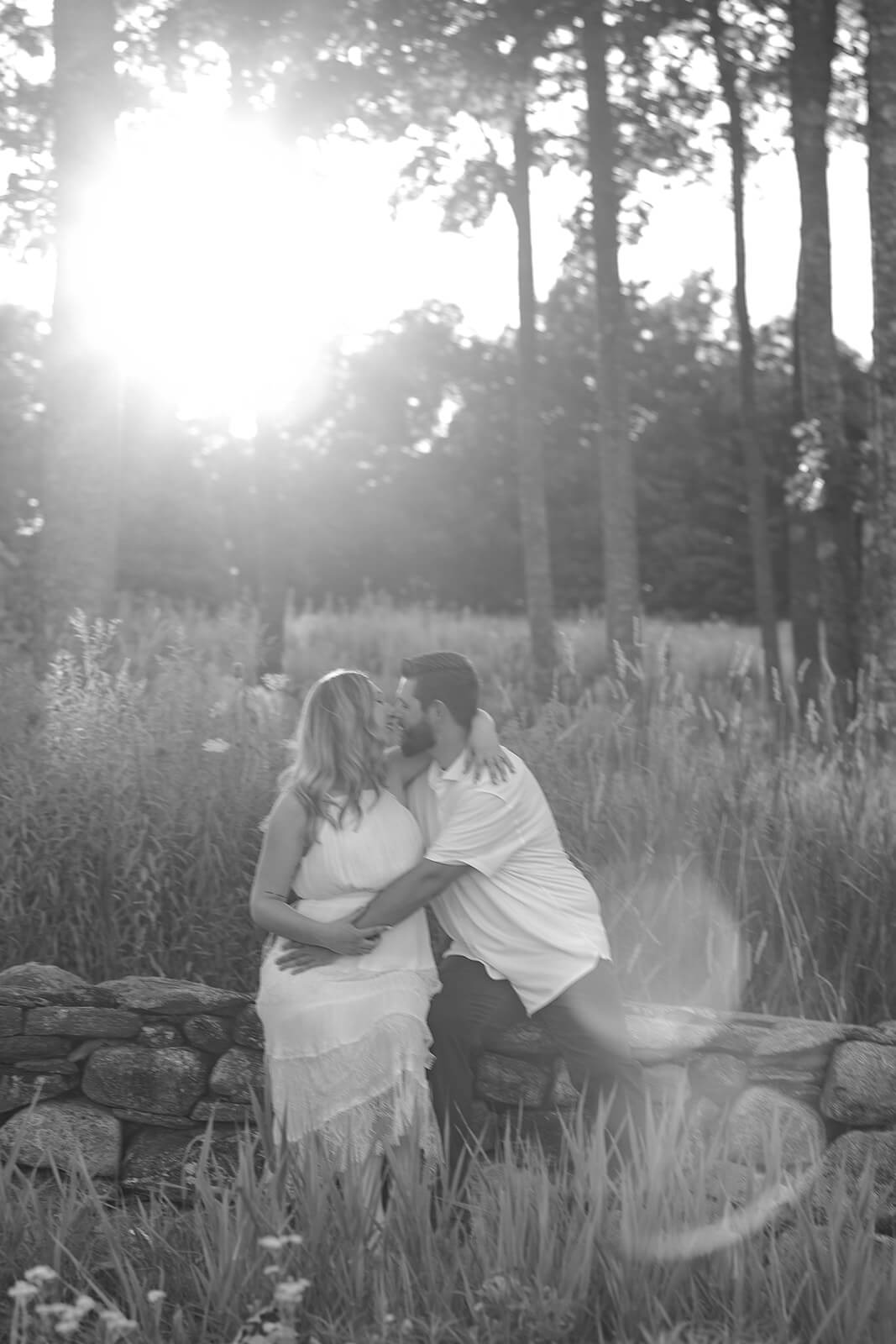 Soft black-and-white portrait of a couple sitting close together in tall grass and facing one another beneath bright sunlight filtering through the trees.