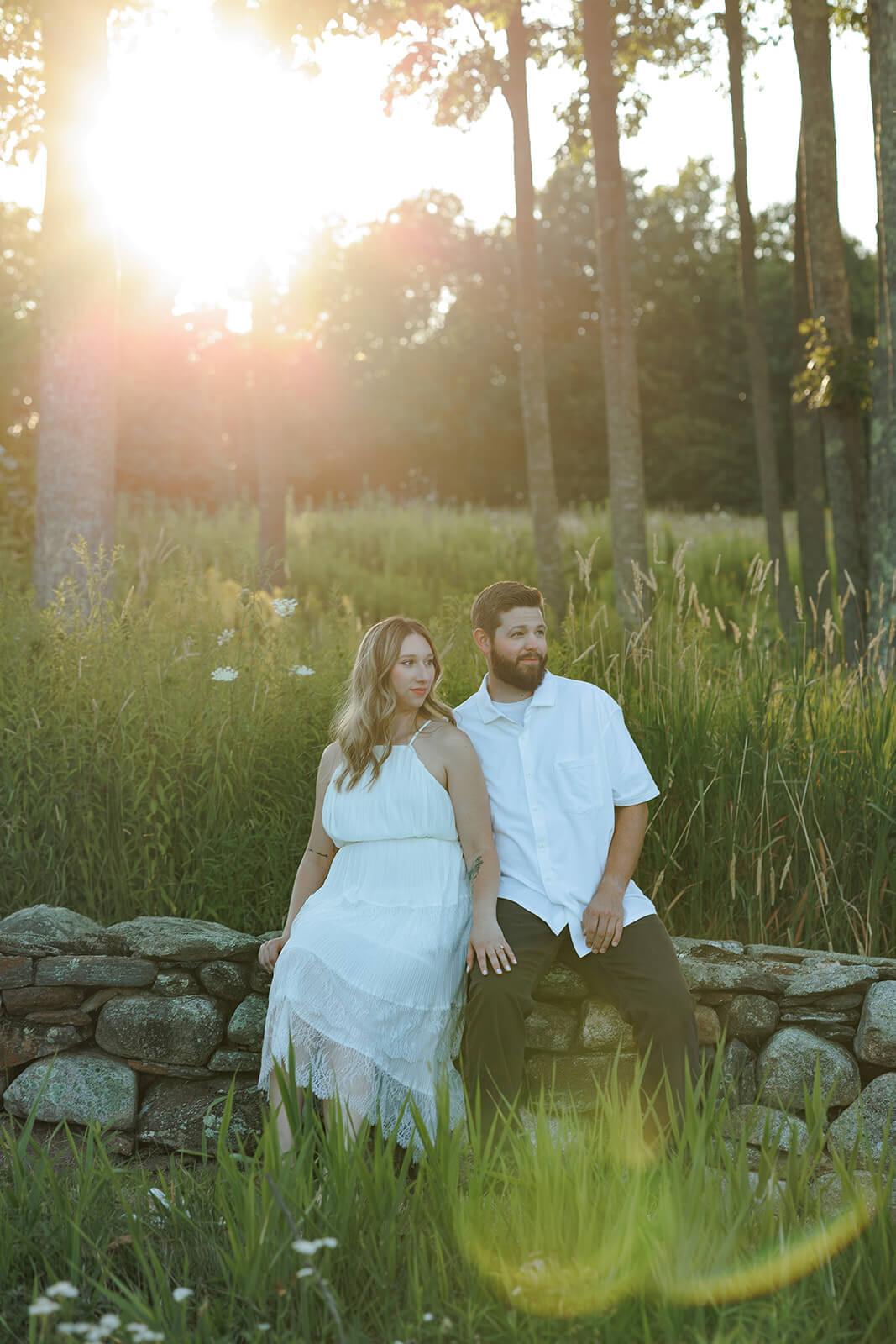 Couple sitting together on a stone wall with tall grass and golden sunlight behind them at Beech Hill Barn.