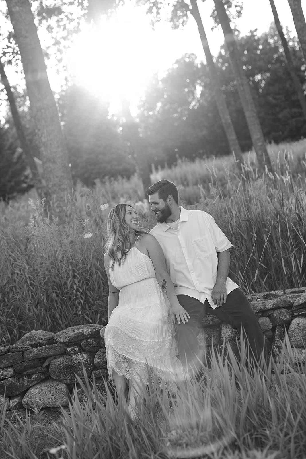 Black-and-white portrait of a couple sitting together on a stone wall and smiling at each other at Beech Hill Barn in Pittston, Maine.