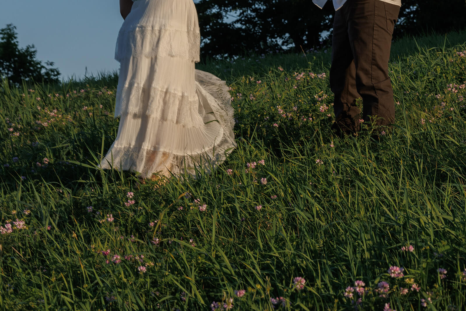 Low-angle cropped photograph of the couple walking through tall grass and pink wildflowers, showing her flowing white skirt and his dark pants.