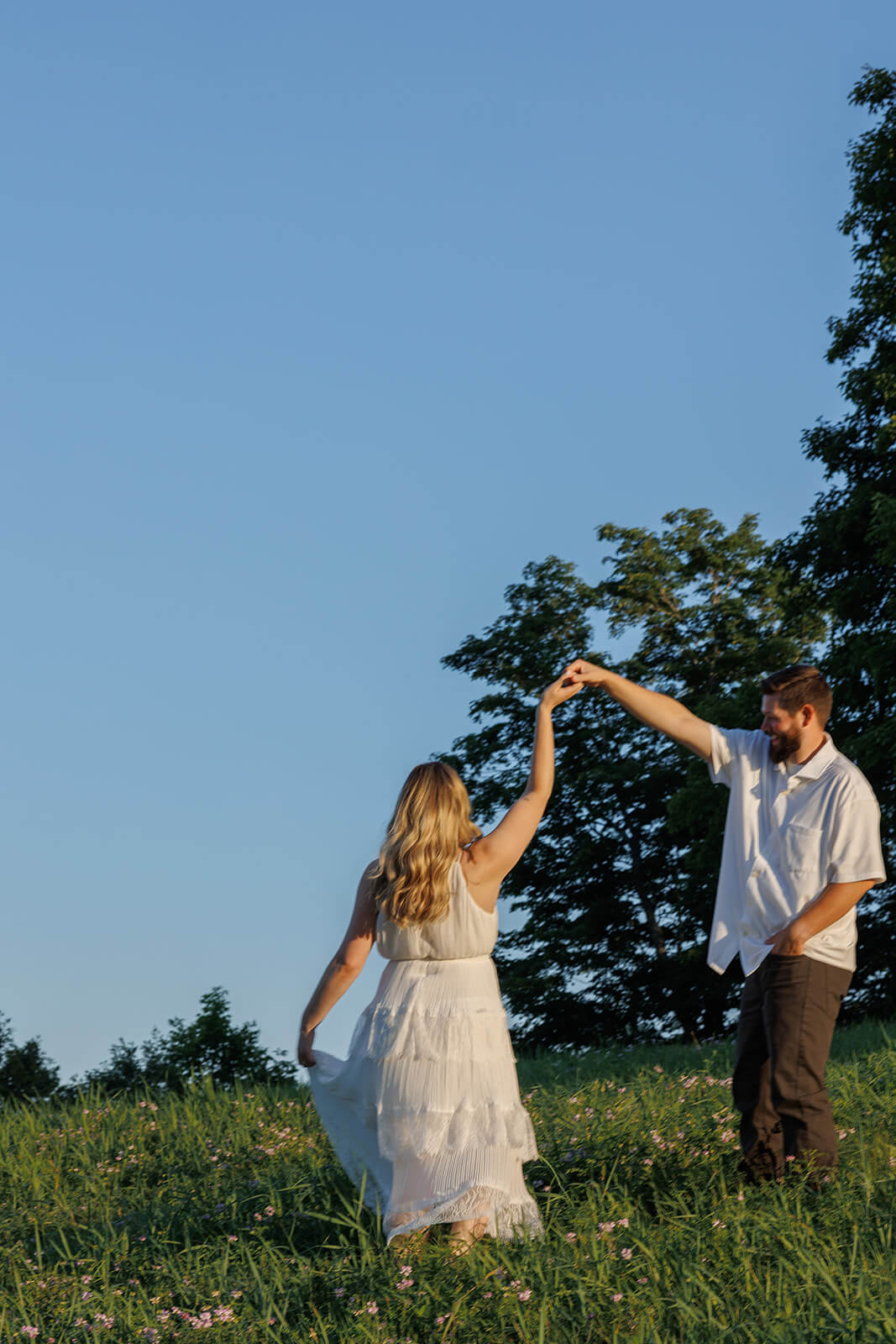 Woman twirling beneath her partner’s raised arm in a green wildflower field beneath a bright blue sky.