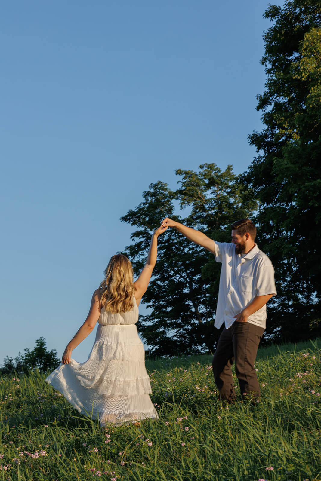 Woman in a white tiered dress twirling beneath her partner’s raised arm in a wildflower field at Beech Hill Barn in Pittston, Maine.