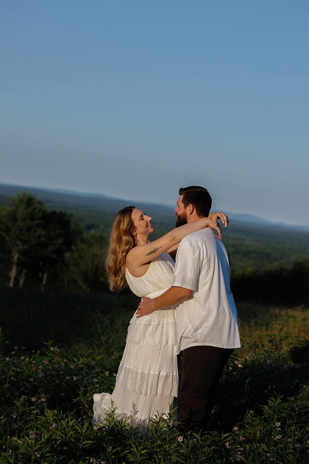 Couple embracing in a hilltop field with distant wooded hills stretching across the horizon at Beech Hill Barn in Pittston, Maine.