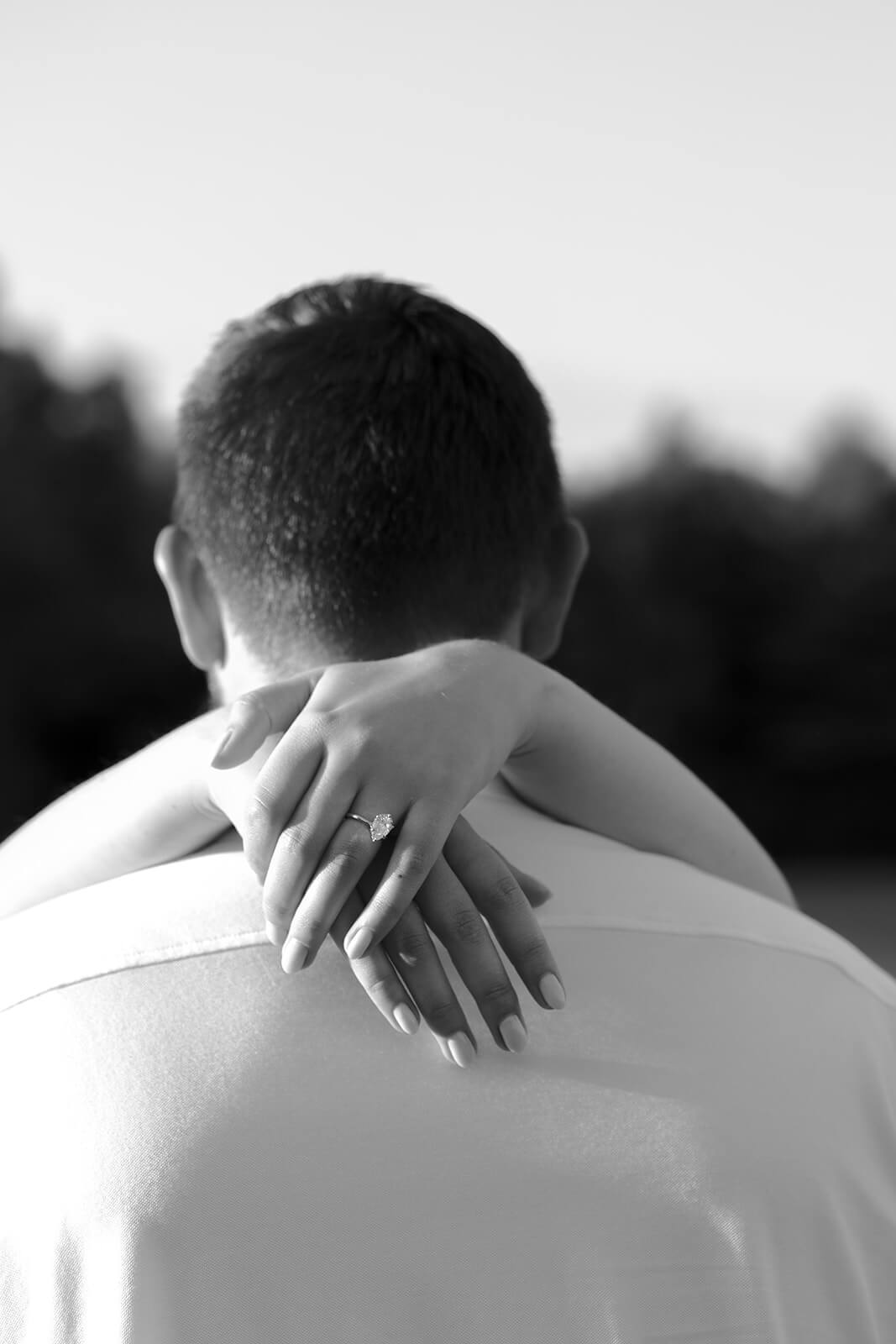 Black-and-white close-up of a woman’s engagement ring as her hand rests around her partner’s shoulders.