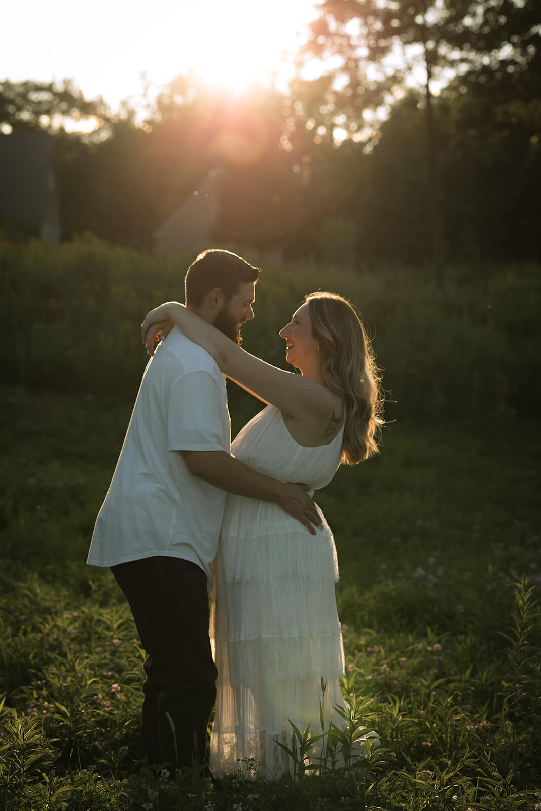 Couple embracing in a wildflower field with the woman’s arms around her partner’s shoulders and golden sunlight behind them at Beech Hill Barn in Pittston, Maine.