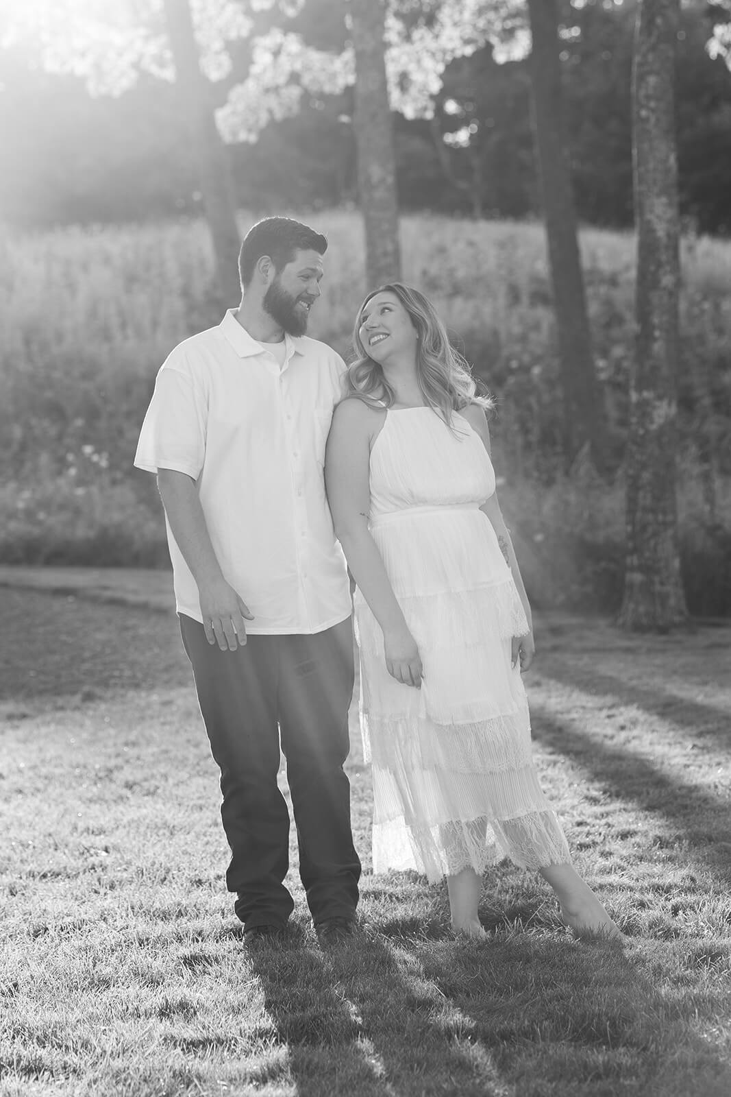 Black-and-white portrait of a couple standing side by side in a field and smiling at each other.
