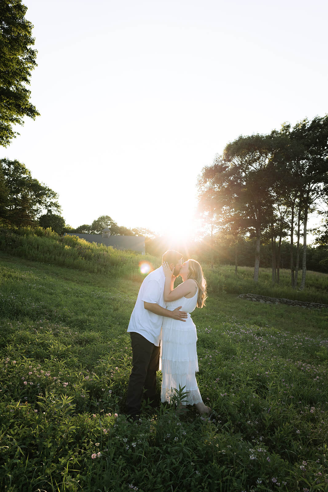 Couple kissing in a green field with the setting sun glowing directly behind them at Beech Hill Barn.