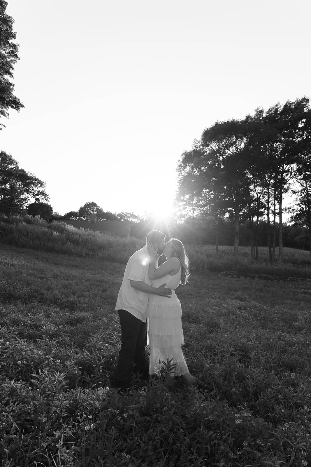 Black-and-white wide portrait of a couple kissing in a grassy field with the low sun shining behind them at Beech Hill Barn.