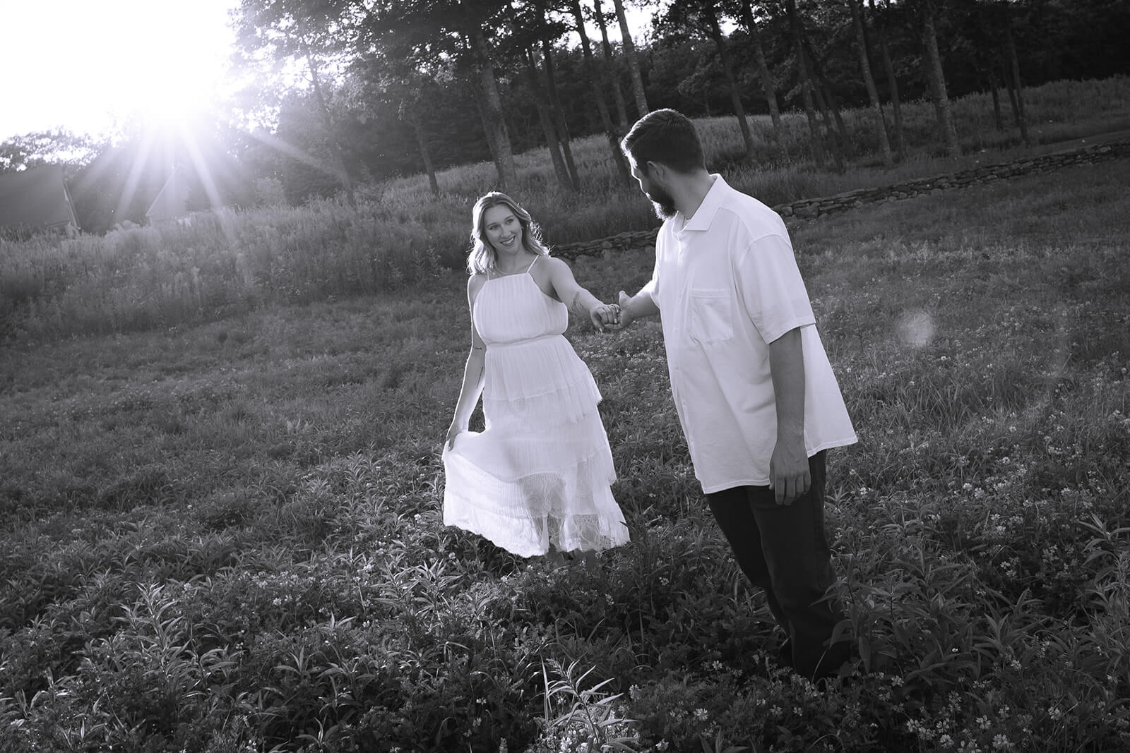 Black-and-white photograph of a woman holding her dress and looking back at her partner as they walk hand in hand through the field at Beech Hill Barn.