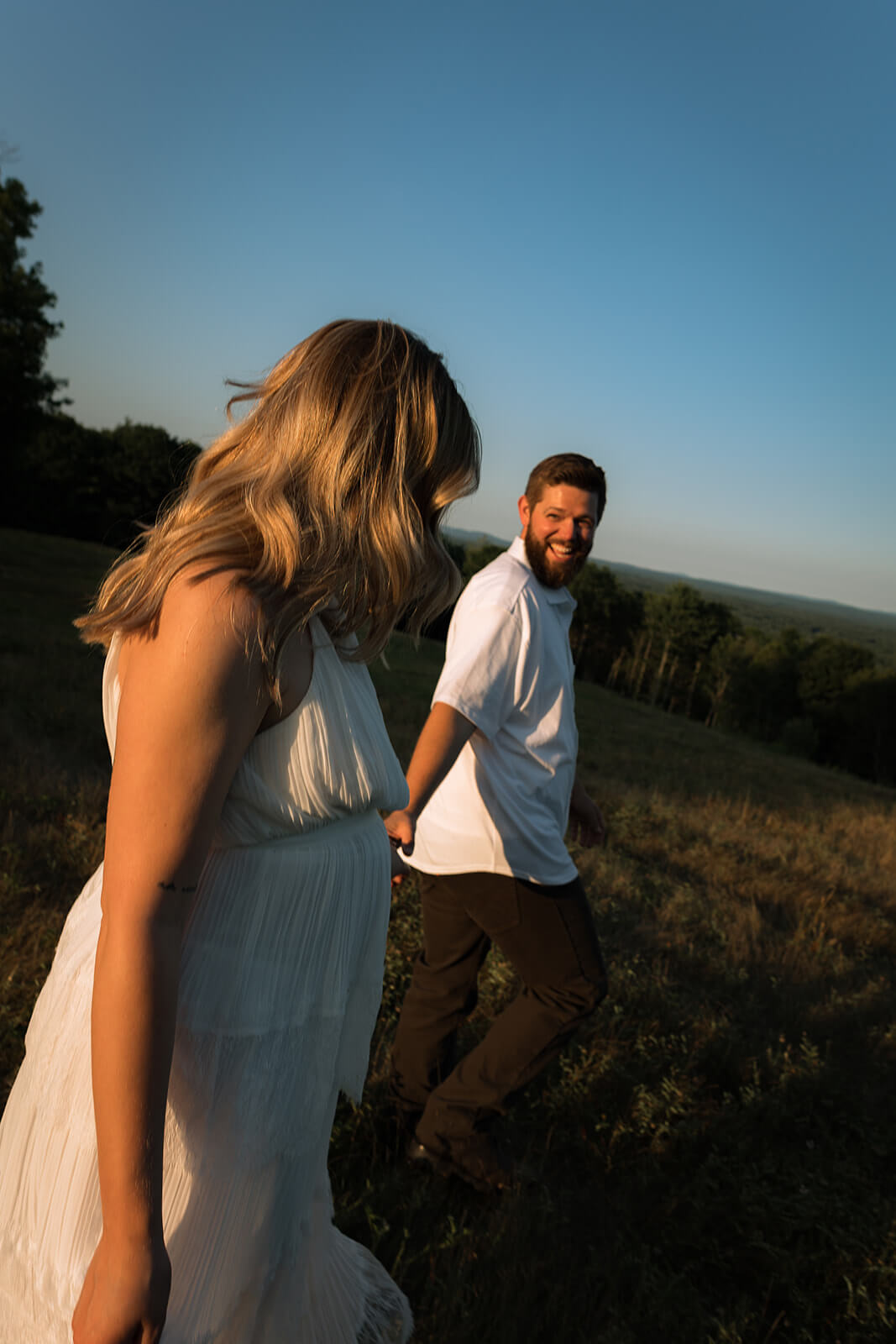 Candid tilted photograph of a woman walking through a hilltop field while her smiling partner follows behind her beneath a clear blue sky.